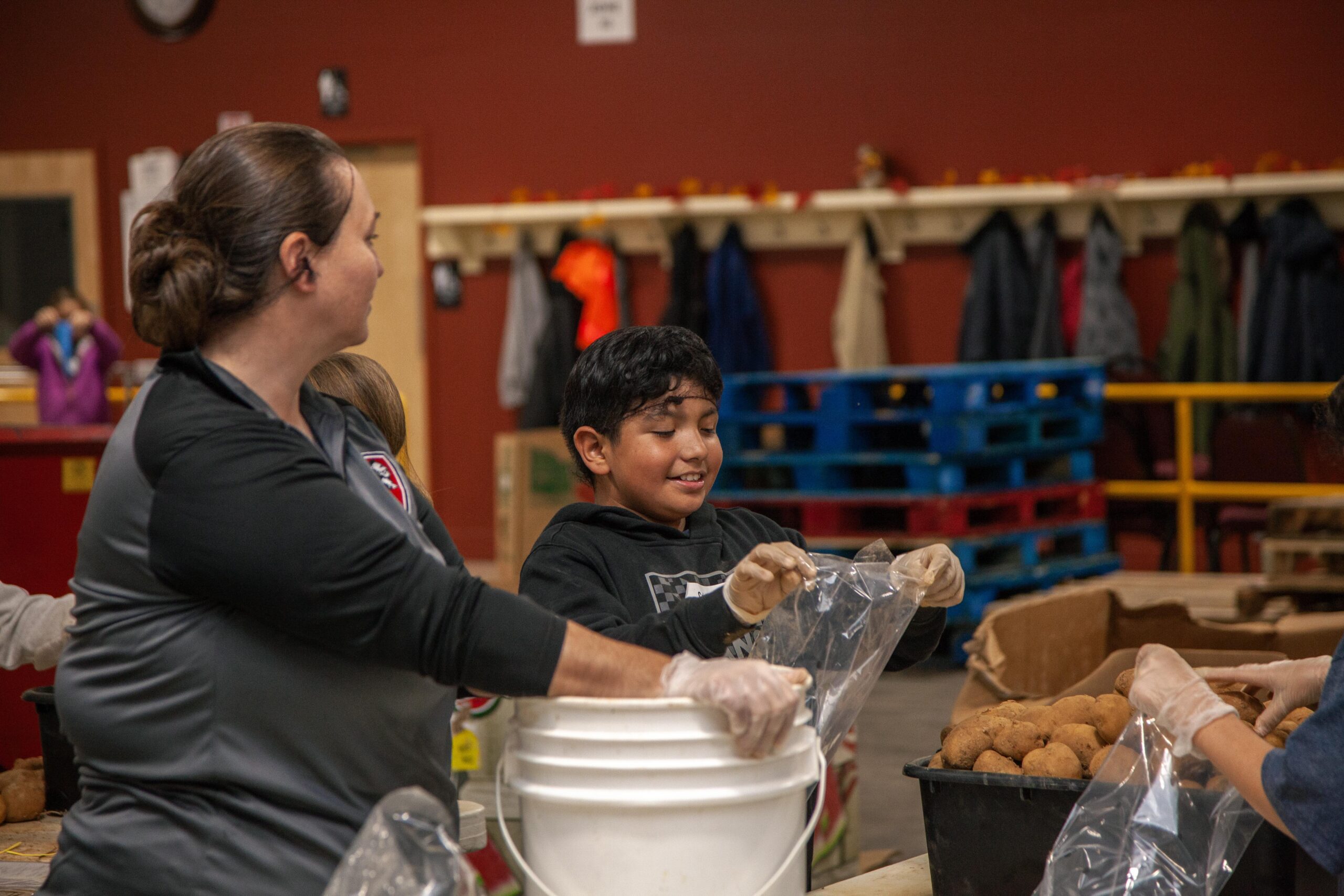 Volunteers packing food items in a community service setting, with a woman and a child smiling while handling plastic bags and food supplies. The background features colorful pallets and coats hanging on a rack, highlighting a warm, collaborative environment.
