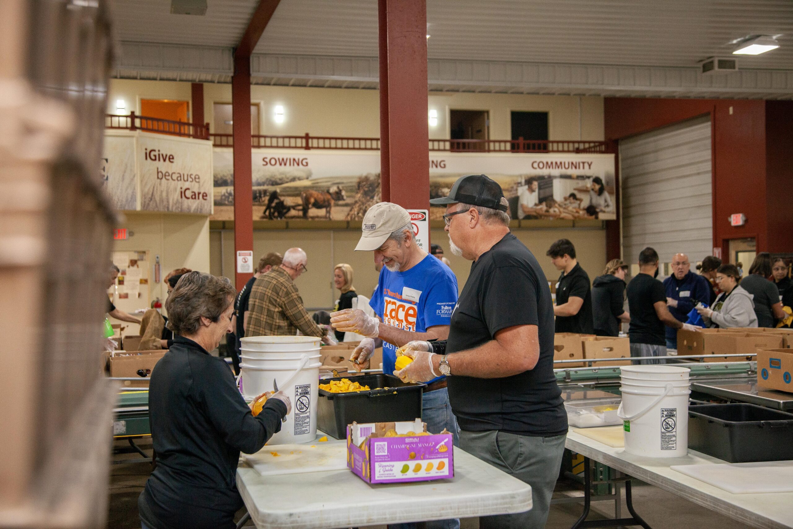 Volunteers participating in a community food sorting event, preparing fruits and vegetables in a large indoor space. The scene shows individuals engaged in food preparation, with various containers and tables set up for efficient organization, promoting community service and collaboration.