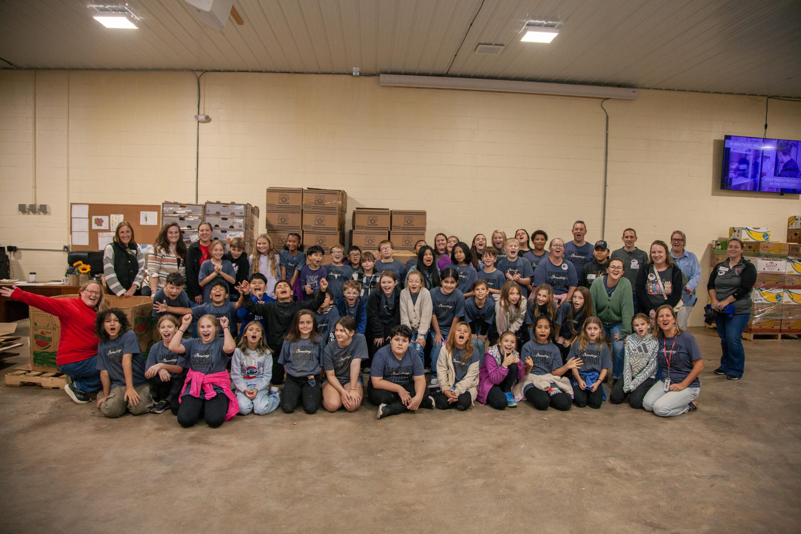 A large group of children and adults smiling and posing for a photo in a food pantry, surrounded by boxes of food and supplies, highlighting community involvement and volunteer efforts.