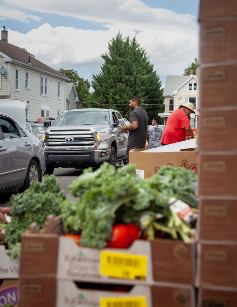 Image of a community food distribution event with fresh vegetables in the foreground, including kale and tomatoes. A man is seen carrying supplies while a silver Toyota truck is parked nearby. Two other individuals are engaged in the activity, set against a backdrop of residential buildings and a clear sky.