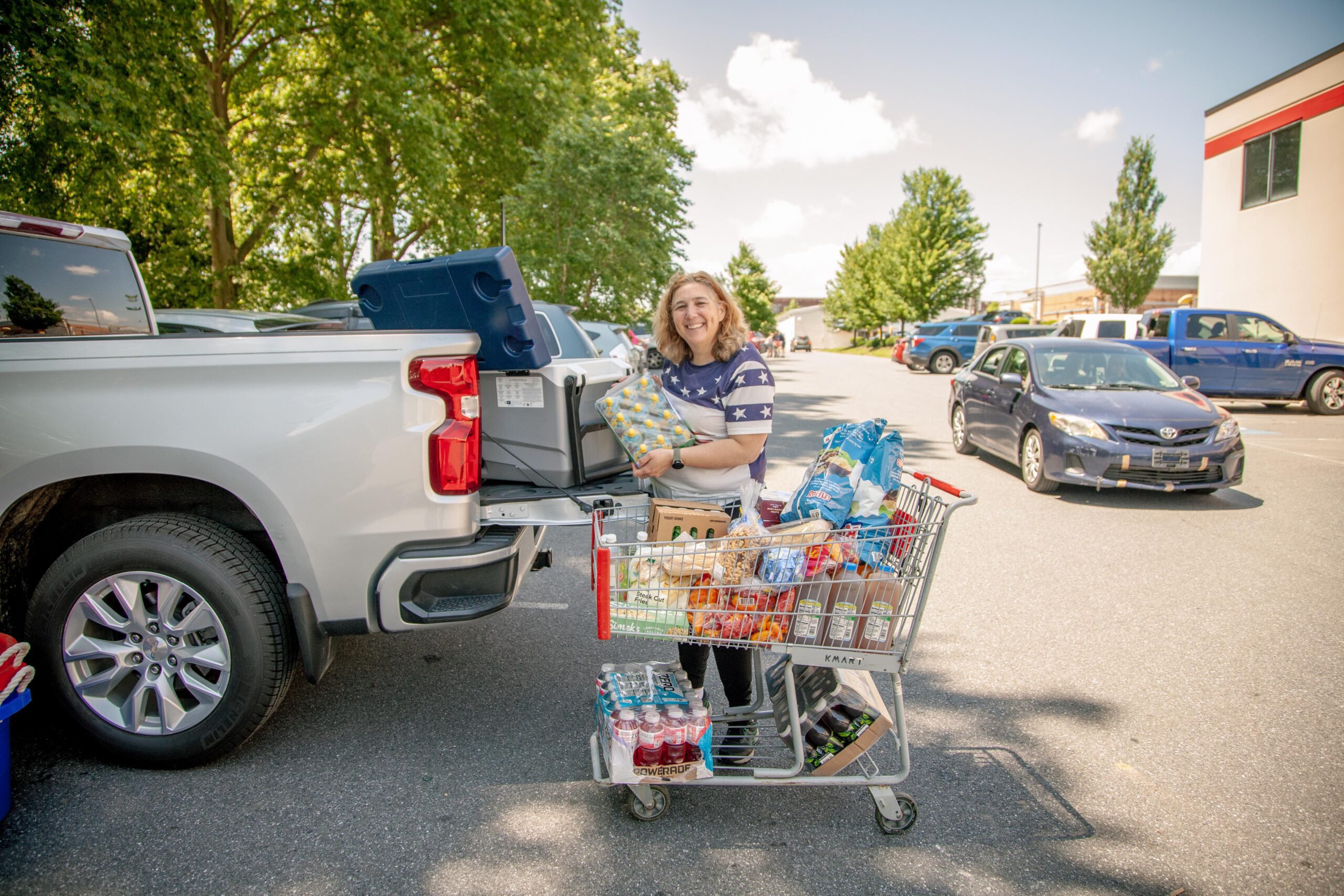 Woman smiling while unloading groceries from a shopping cart into the back of a pickup truck in a parking lot, surrounded by trees and parked cars. The cart is filled with various food items and beverages.