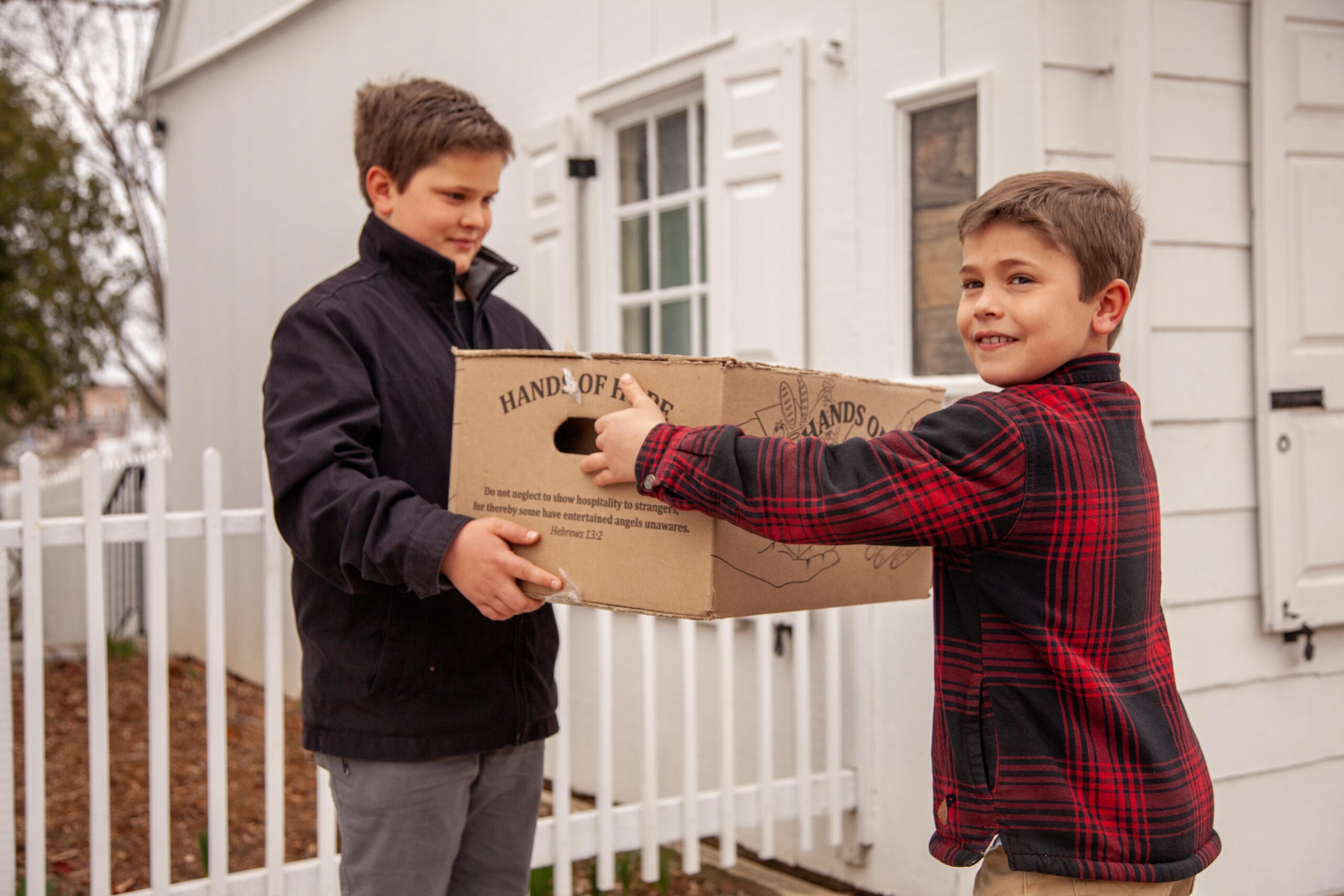 Two boys are participating in a community service activity, one handing a cardboard box labeled "Hands of Hope" to the other. The scene takes place outside a house with a white picket fence, showcasing themes of generosity and kindness.