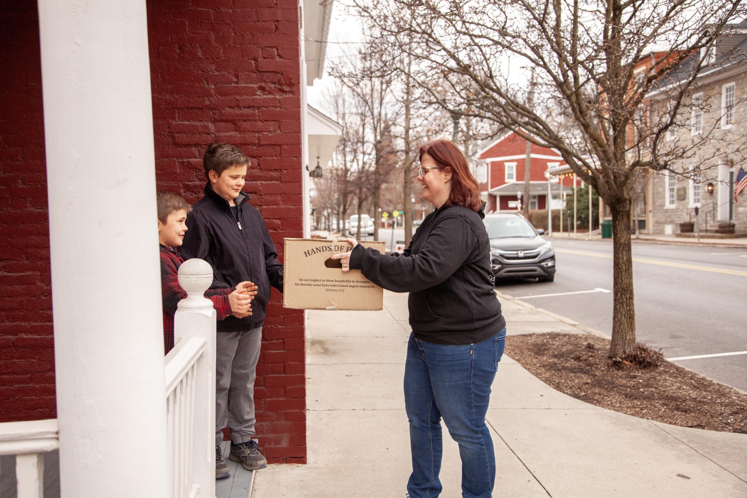 Woman handing a cardboard box to two boys on a sidewalk in a small town, showcasing community support and kindness during a chilly day.