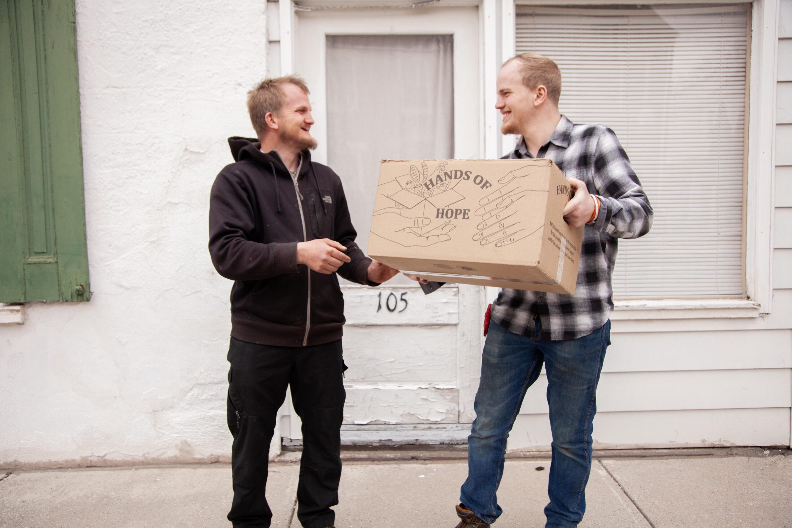 Two men smiling and exchanging a box labeled "Hands of Hope" in front of a house, symbolizing community support and charitable giving.