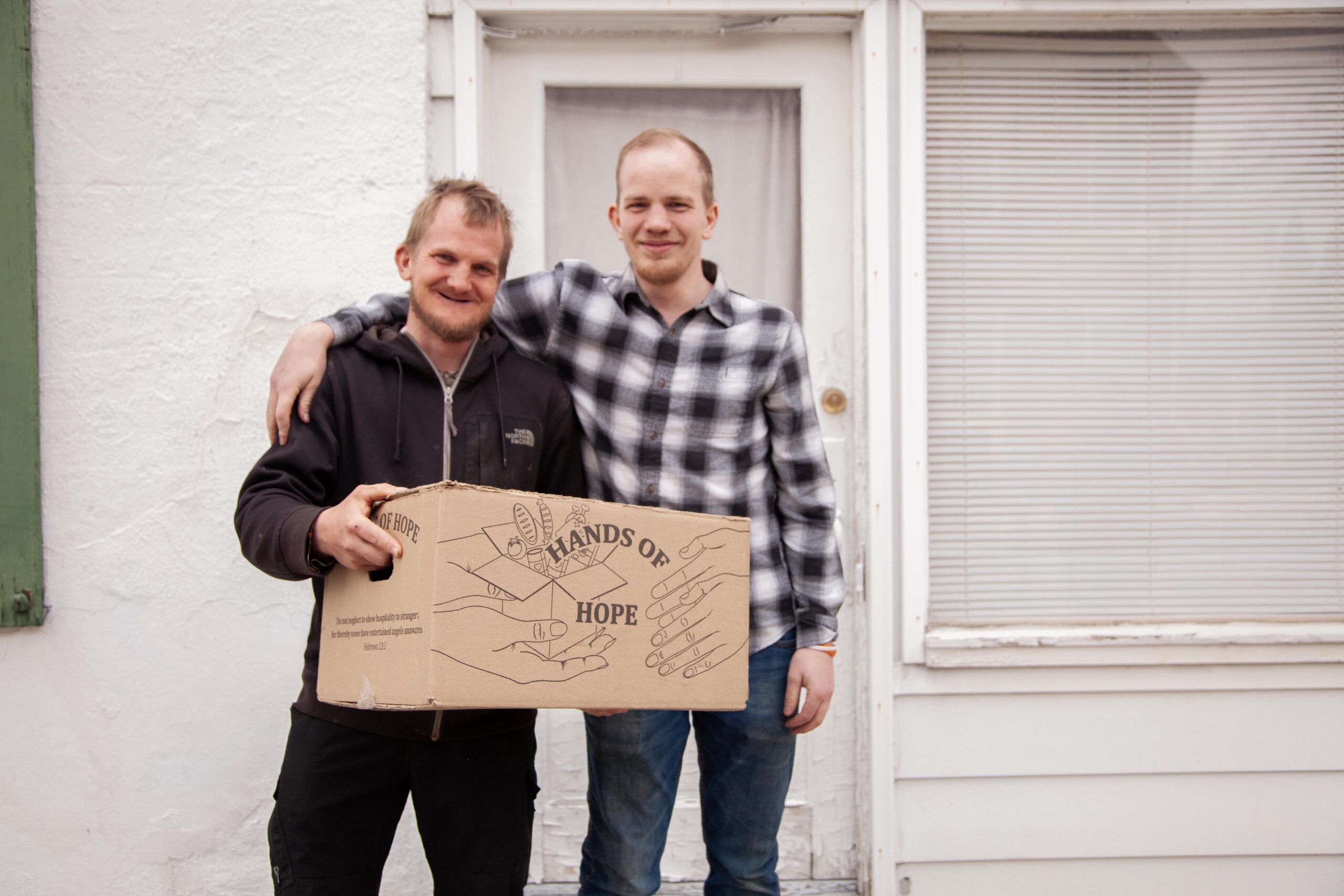 Two men standing together outside a house, holding a cardboard box labeled "Hands of Hope." The background features a white wall and a door with blinds. The men are smiling, showcasing a spirit of community and support.