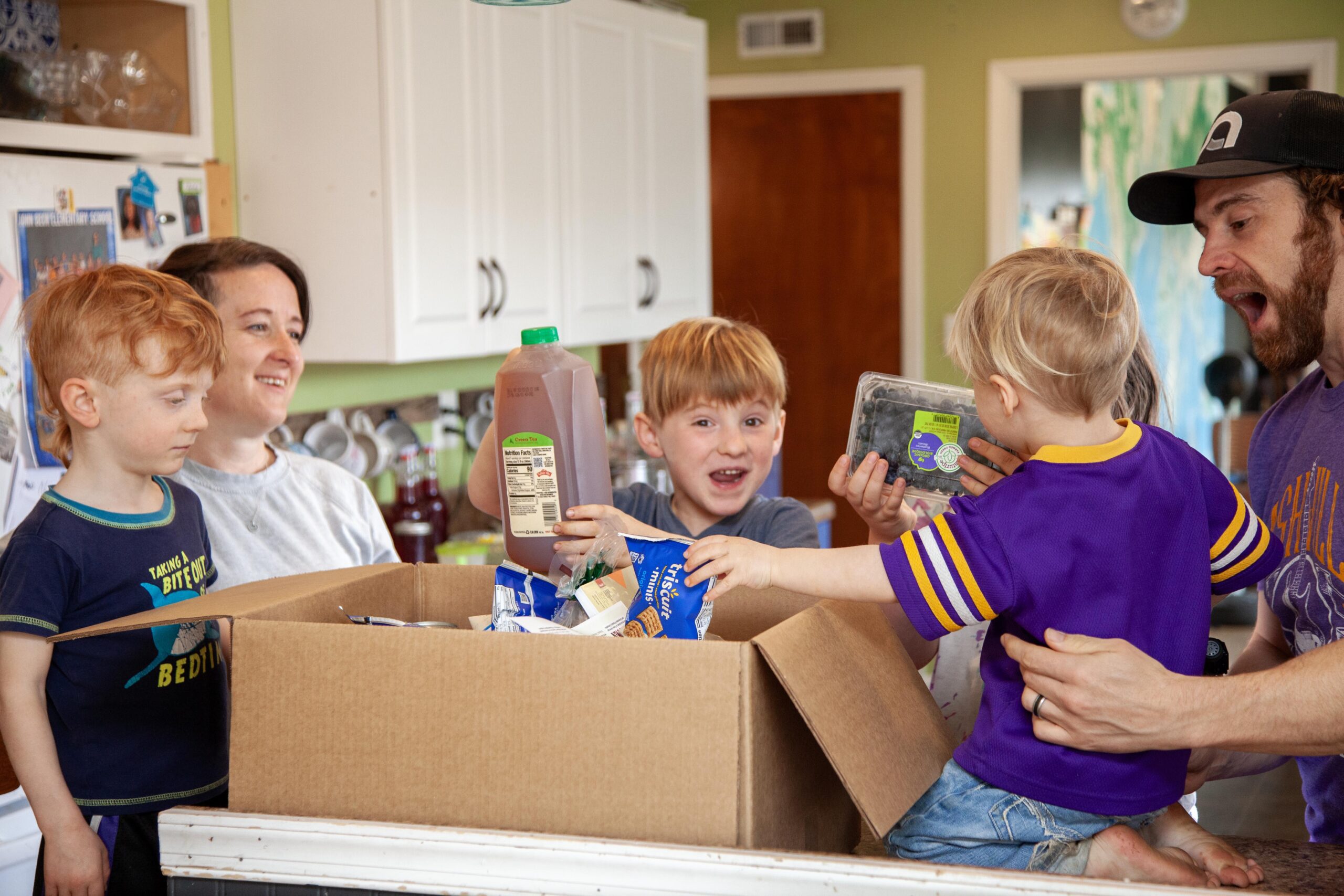 A family excitedly unpacks a large box of groceries in a bright kitchen, with two young boys eagerly holding items like juice and blueberries, while a woman smiles nearby and a man assists the children.