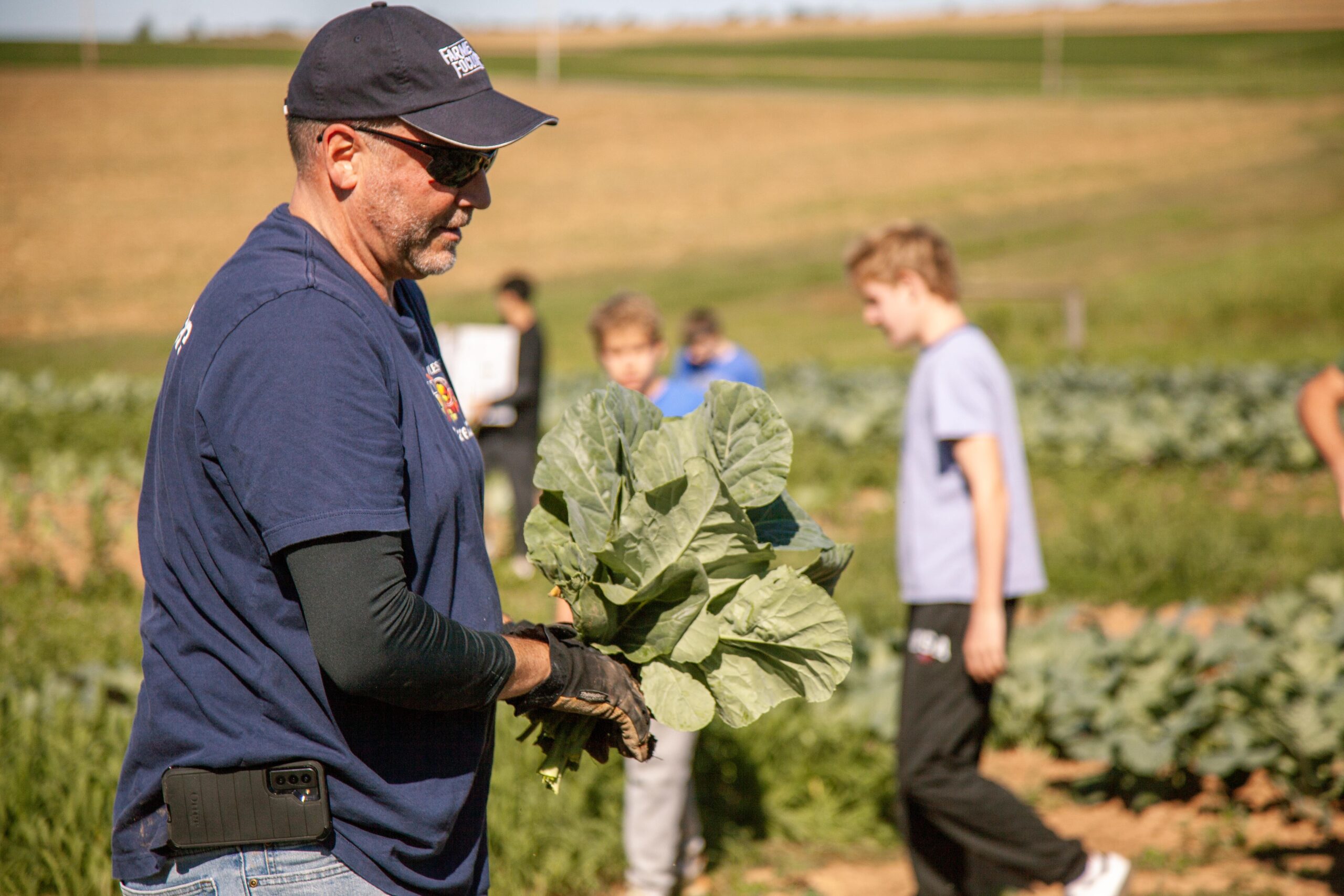 A farmer wearing sunglasses and gloves holds a bundle of fresh greens while a group of young volunteers work in the background on a sunny day in a vegetable field. The image captures the essence of agricultural teamwork and sustainable farming practices.