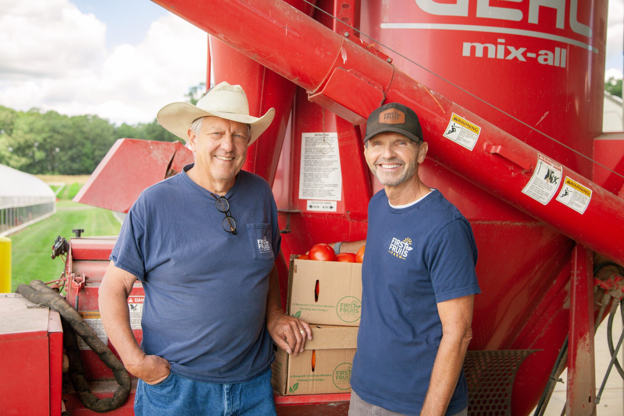 Two farmers smiling beside a red agricultural mixer, with boxes of freshly harvested tomatoes in the background, showcasing a community-focused farming operation.