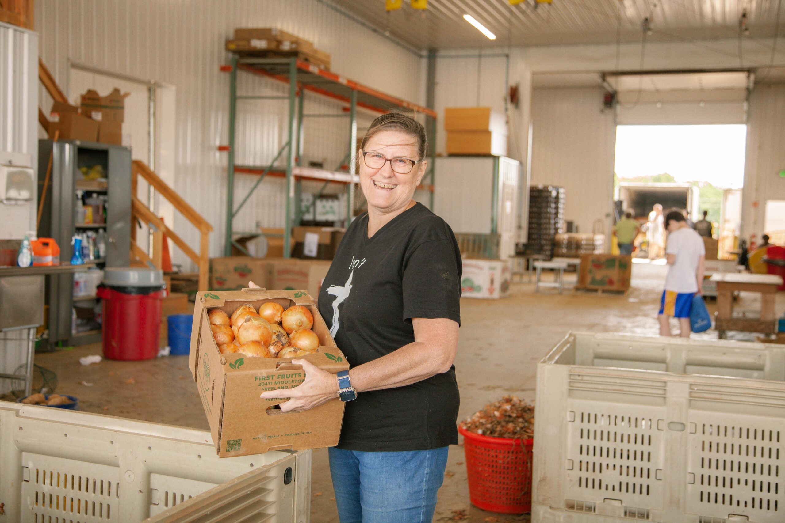 Woman smiling while holding a box of freshly harvested onions in a produce warehouse, with various storage containers and other workers in the background.