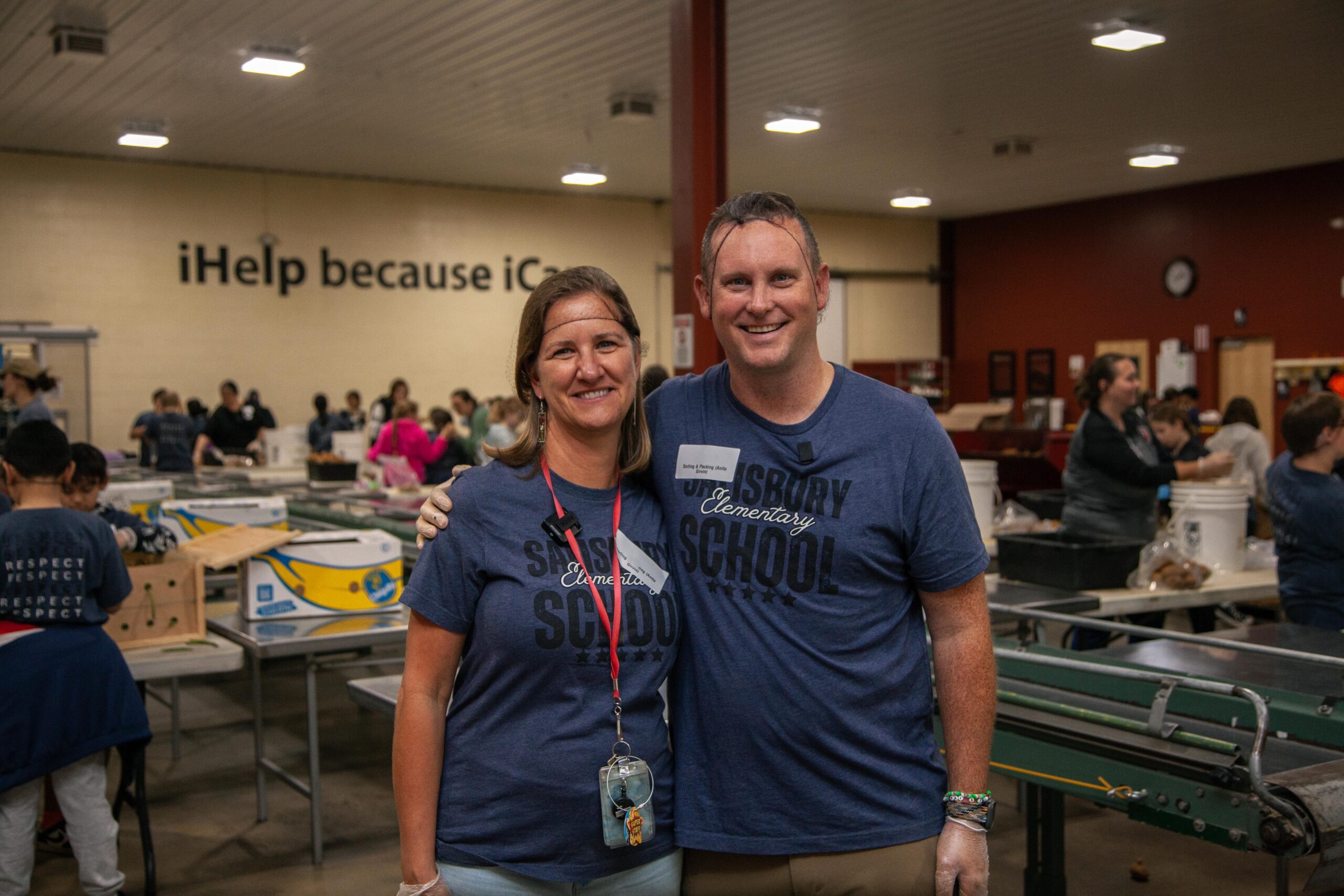 Volunteers smiling at a community service event in a school cafeteria, wearing matching shirts that say "Salisbury Elementary School." The background features a wall with the phrase "iHelp because iCan," while other volunteers work on various tasks at tables filled with supplies.