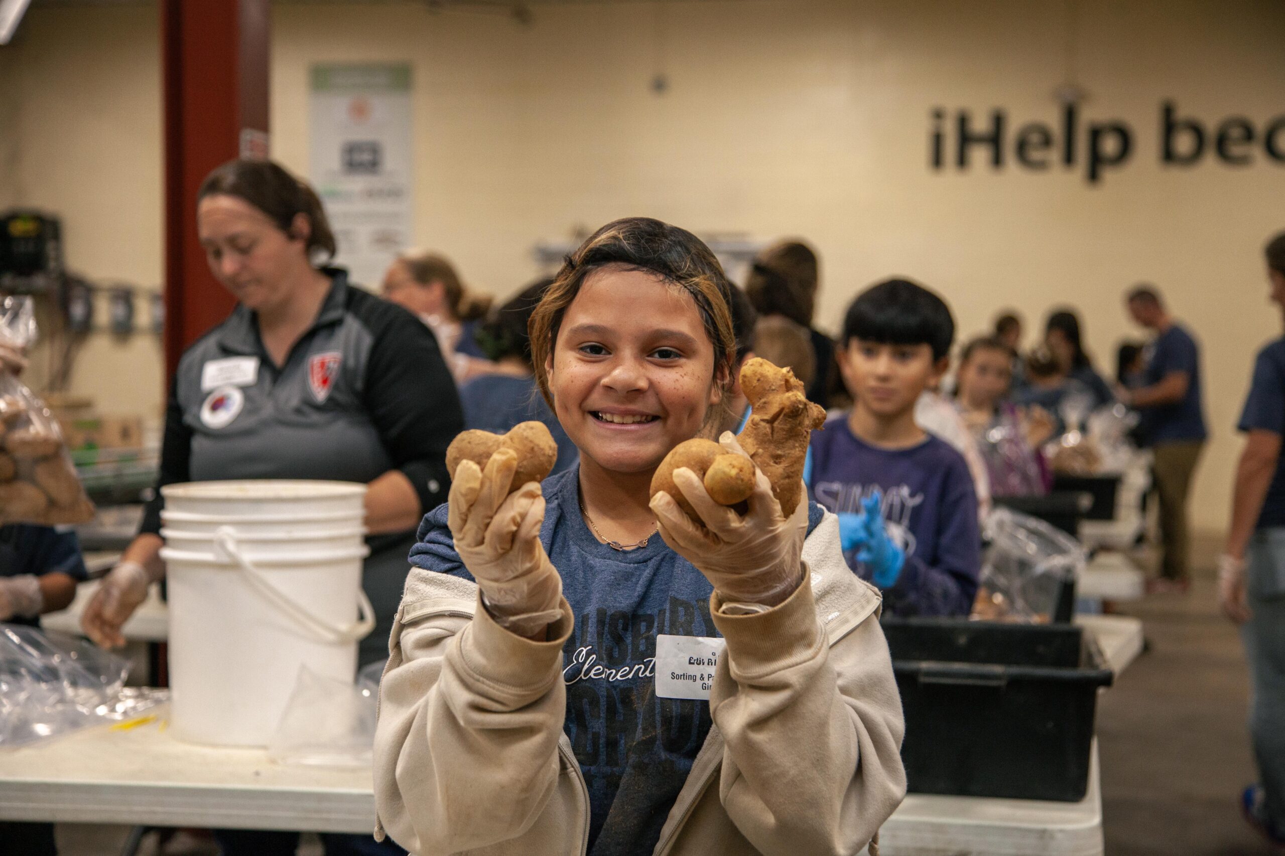Child smiling while holding potatoes in a food sorting event, with volunteers in the background preparing food for distribution.