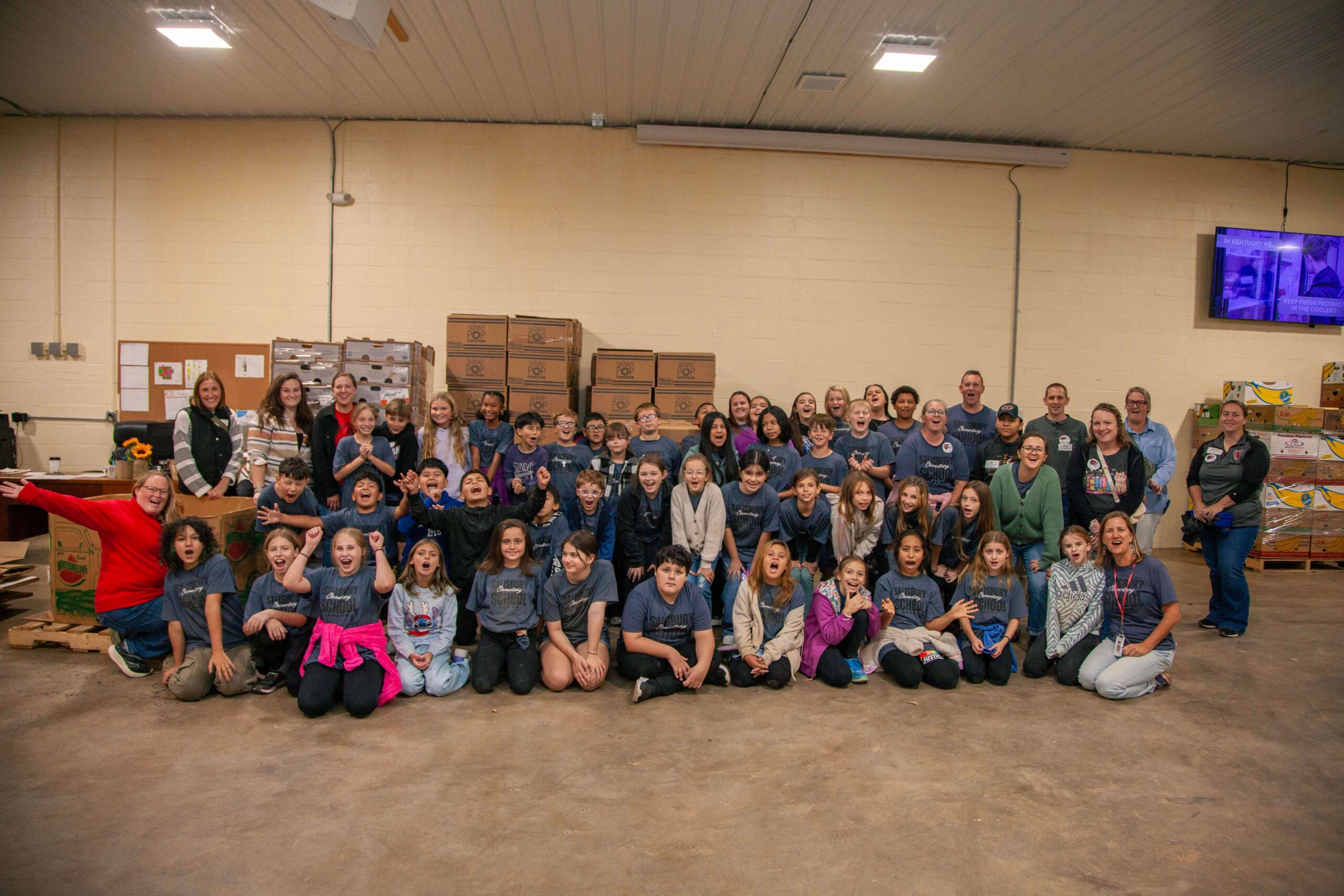 Group photo of students and volunteers at a community food bank event, smiling and celebrating their contribution to helping those in need. Boxes of food are stacked in the background, showcasing the organization's efforts in food distribution.
