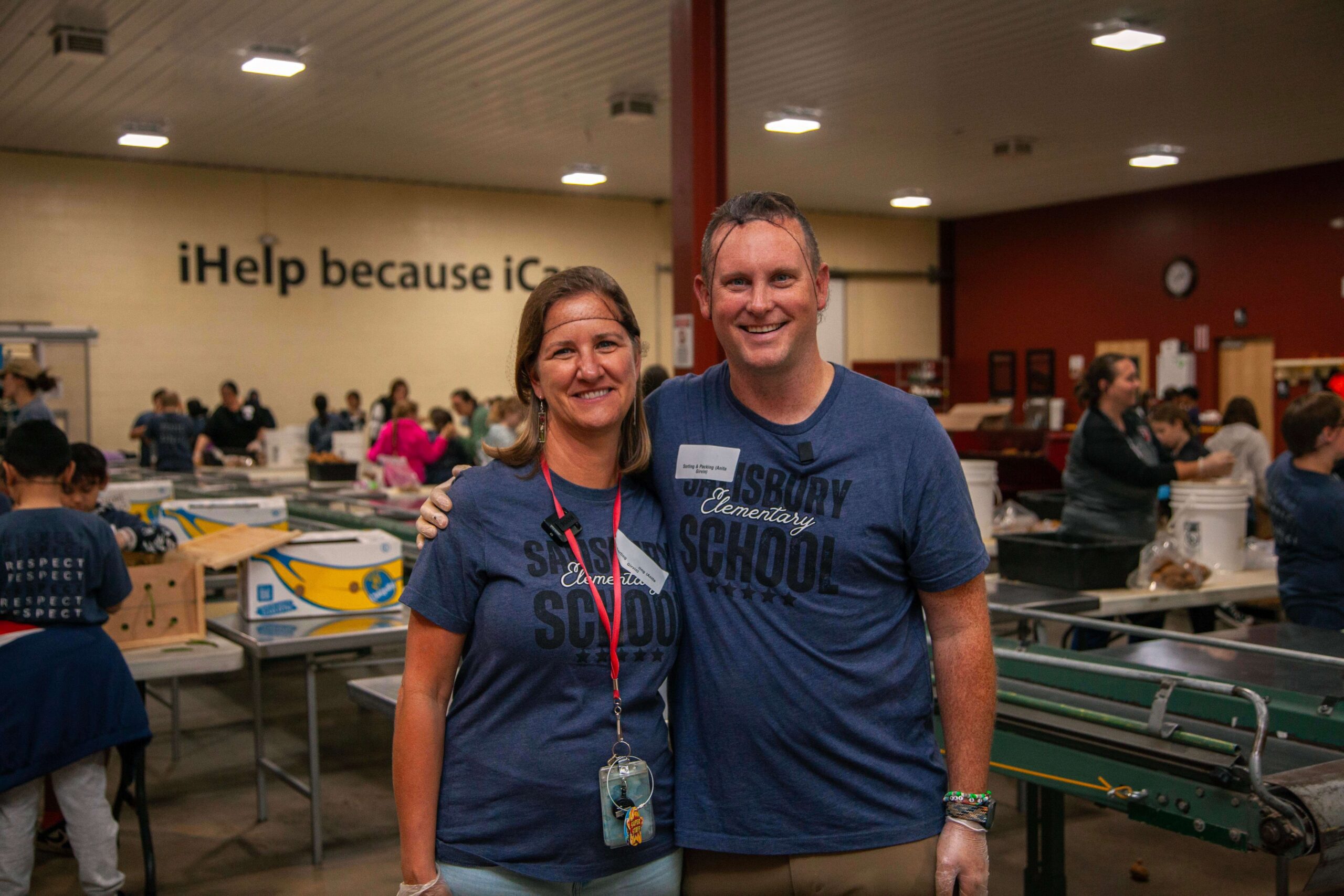 Two volunteers from Salisbury Elementary School smile for a photo while participating in a community service event, with other volunteers working in the background. The setting features tables filled with supplies and a wall displaying the message "iHelp because iCan."