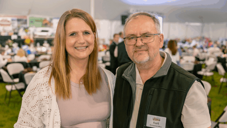 A smiling woman and man pose together at a formal event held under a tent, with tables and decorations visible in the background. The man is wearing glasses and a name tag indicating his role as a Development Officer.