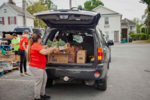 A woman in a red shirt loads fresh produce and boxes into the back of an SUV at a community food distribution event, with volunteers and parked vehicles in the background.