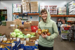 Teen volunteer smiling while holding fresh produce in a busy food pantry, surrounded by boxes of fruits and vegetables.