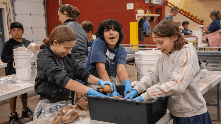Children volunteering in a community food distribution center, sorting and packing potatoes into bags while wearing gloves. The scene captures teamwork and the spirit of giving back, with adults supervising in the background.