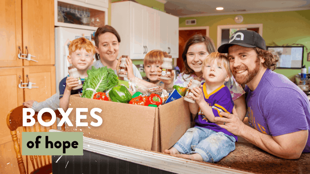 A happy family of five poses in a kitchen, showcasing a large cardboard box filled with fresh produce and healthy snacks, symbolizing community support through the "Boxes of Hope" initiative.