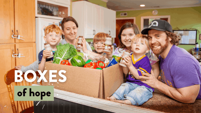 A happy family of five poses in a kitchen, showcasing a large cardboard box filled with fresh produce and healthy snacks, symbolizing community support through the "Boxes of Hope" initiative.
