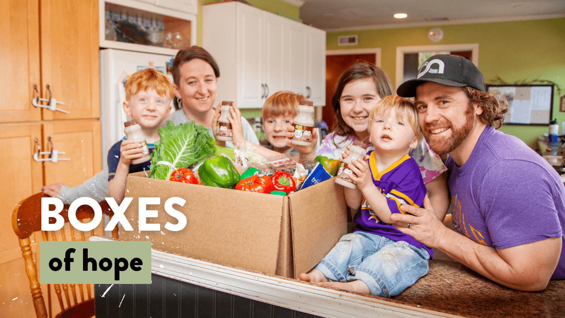 A happy family of five poses in a kitchen, showcasing a large cardboard box filled with fresh produce and healthy snacks, symbolizing community support through the "Boxes of Hope" initiative.