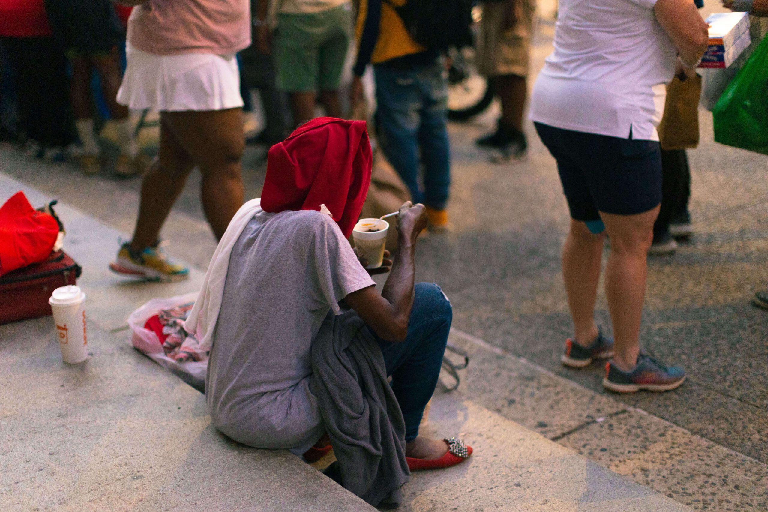 A person sitting on steps, wearing a red hood and gray clothing, holding a cup, while people walk by in a busy urban setting.