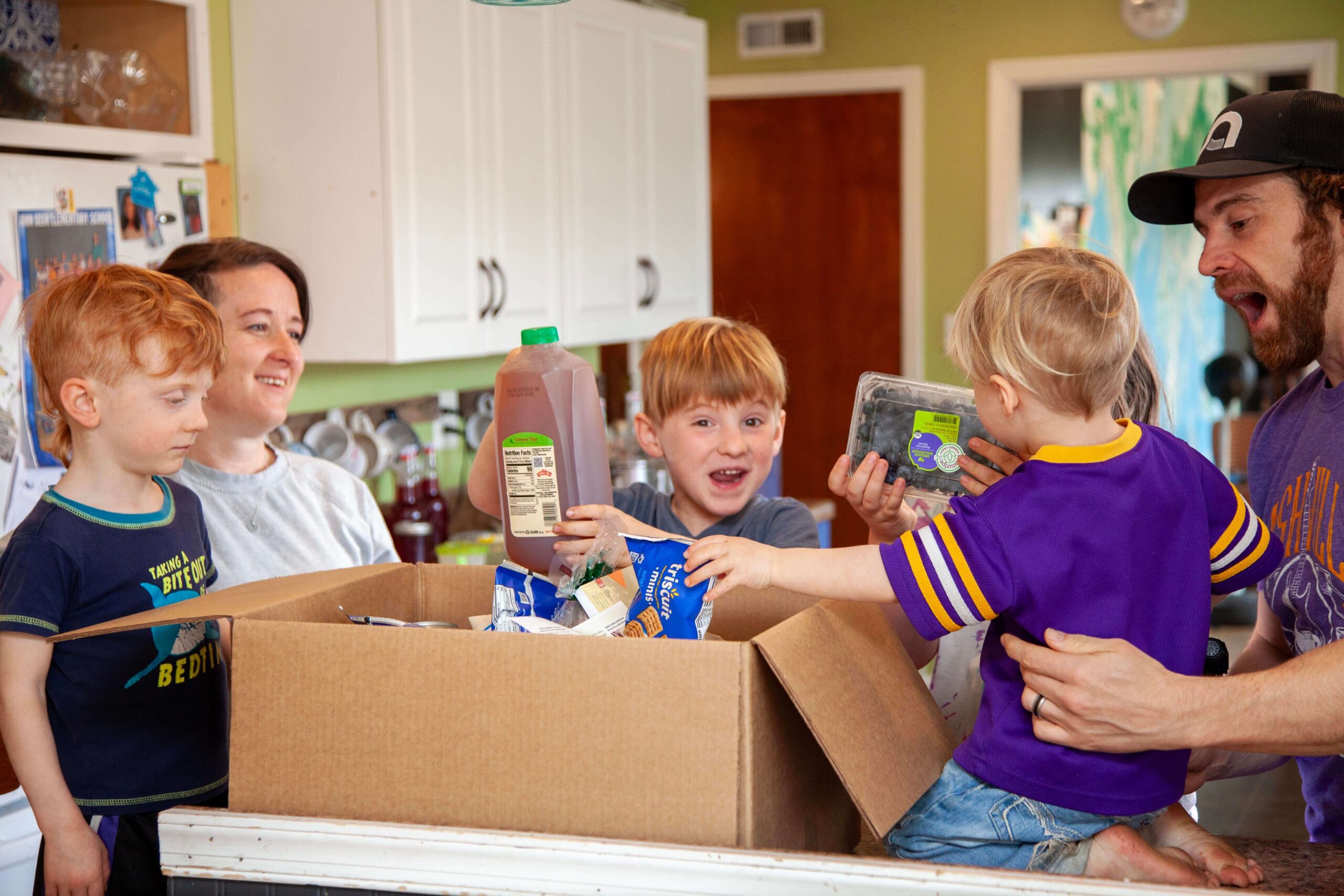 Family excitedly unpacking a cardboard box filled with groceries in a bright kitchen, featuring children holding juice and blueberries while adults smile and engage.