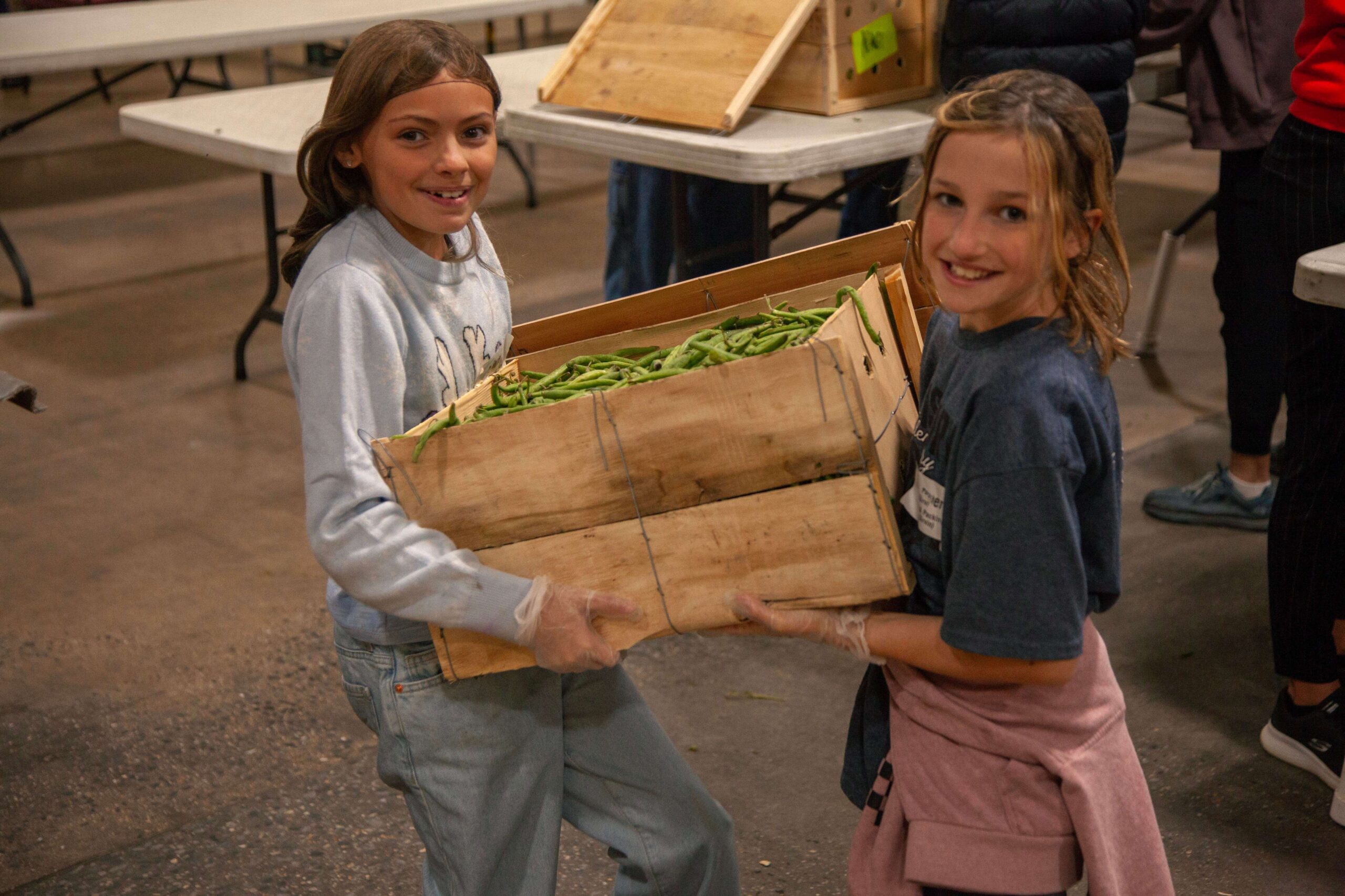 Two children smiling while carrying a wooden crate filled with green beans in a community setting, surrounded by tables and other volunteers.