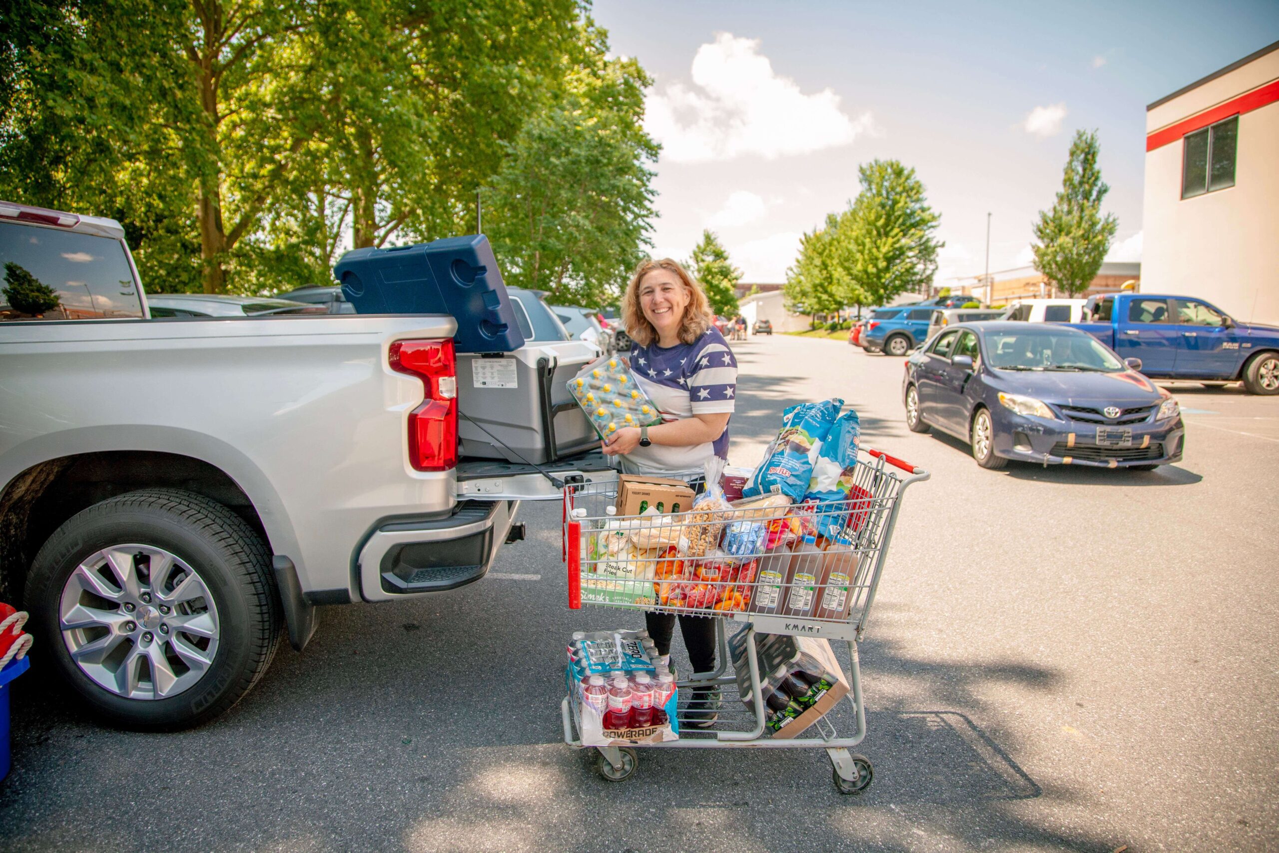 Smiling woman unloading groceries from a shopping cart into the back of a pickup truck in a parking lot, surrounded by trees and parked cars on a sunny day.