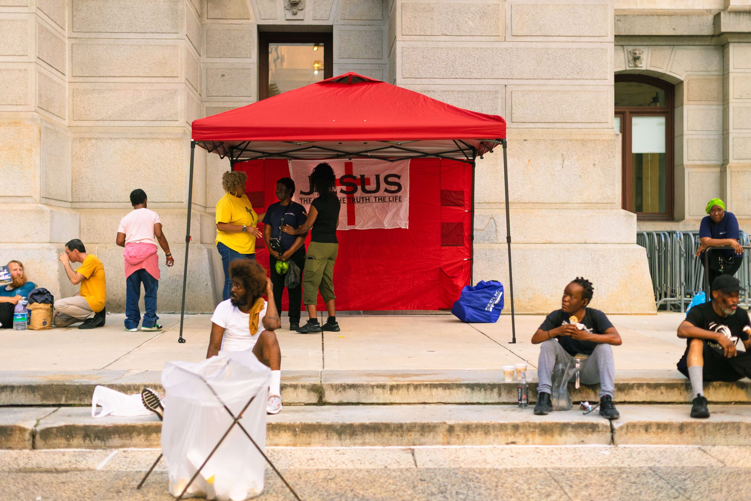 Community gathering under a red tent featuring a religious banner, with diverse individuals engaged in conversation and sitting on the steps of a public building. The scene captures a moment of interaction and support among attendees.