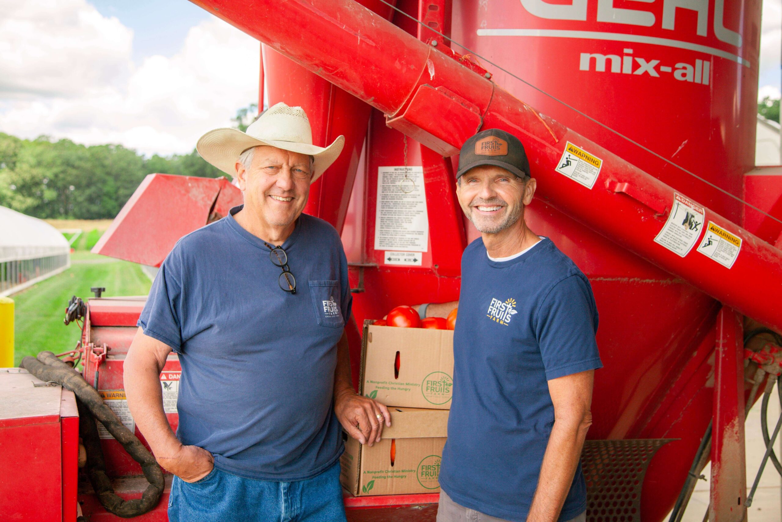 Two farmers standing in front of a red agricultural mixer, smiling and posing for the camera. They are wearing matching navy blue shirts with a logo and a cowboy hat. Behind them, there are boxes of fresh produce, likely tomatoes, and a greenhouse visible in the background, indicating a productive farming environment.