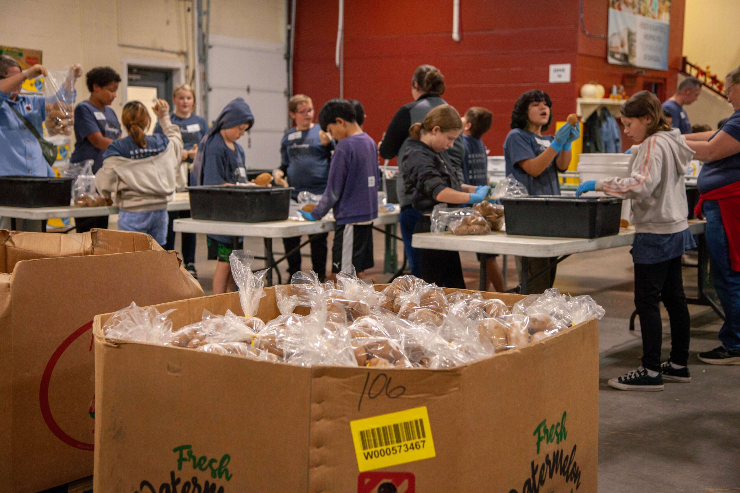 Volunteers packing food items in a community center, with bags of baked goods in foreground and children helping at tables in the background.