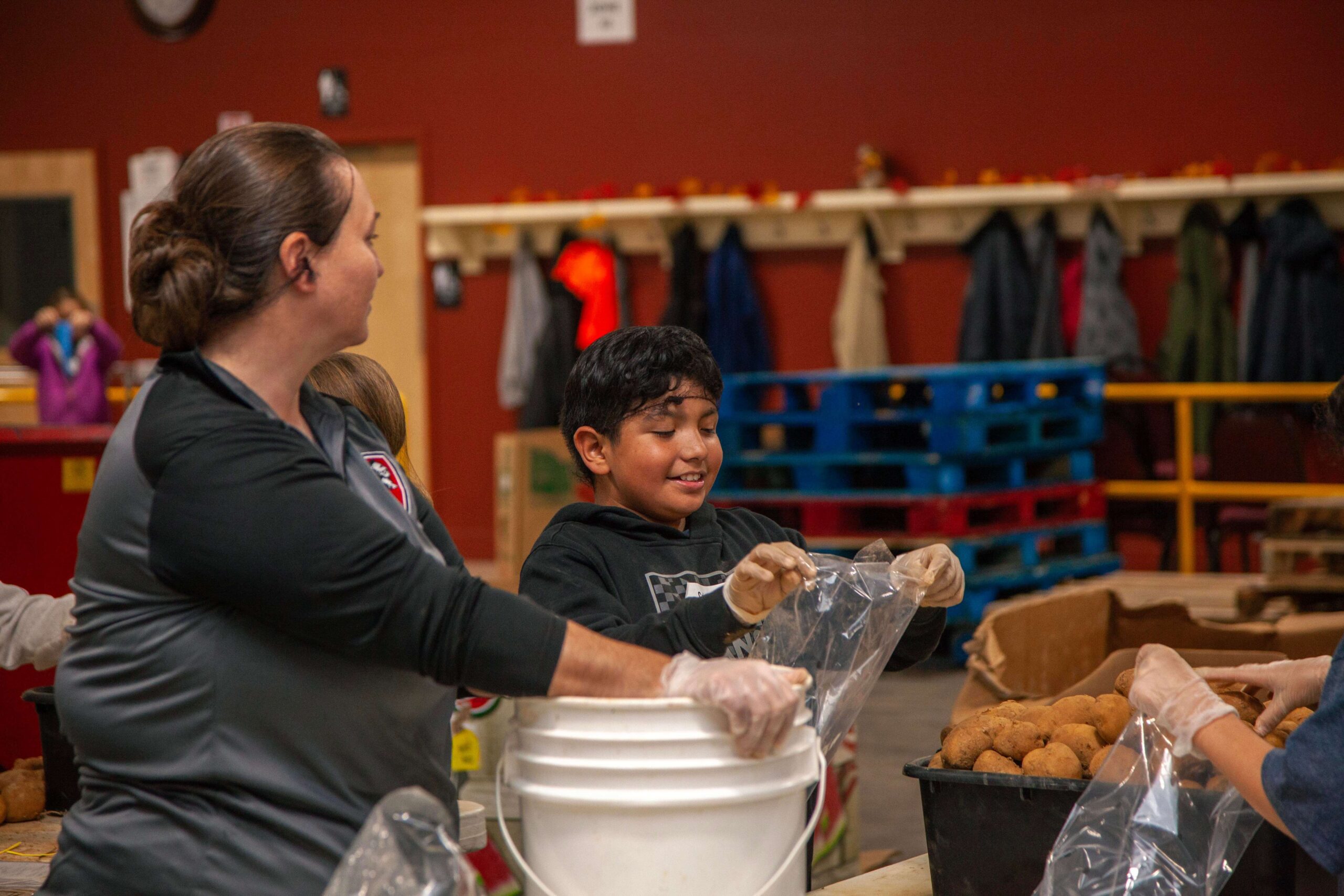 Volunteers packaging food items in a community center, with a woman assisting a child in placing items into plastic bags. The scene highlights teamwork and community service efforts.