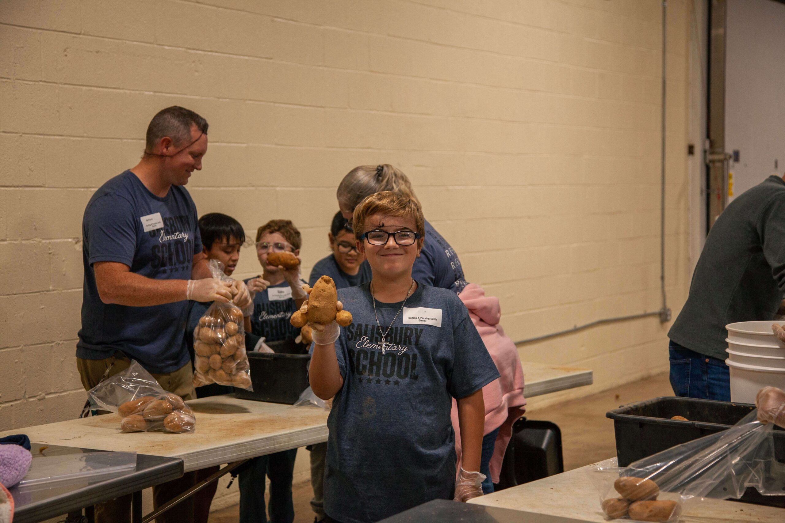 Students and volunteers at Salisbury Elementary School participate in a community service event, sorting and packing sweet potatoes in a large indoor space. A young boy in a blue shirt proudly holds a sweet potato while others assist in the background.