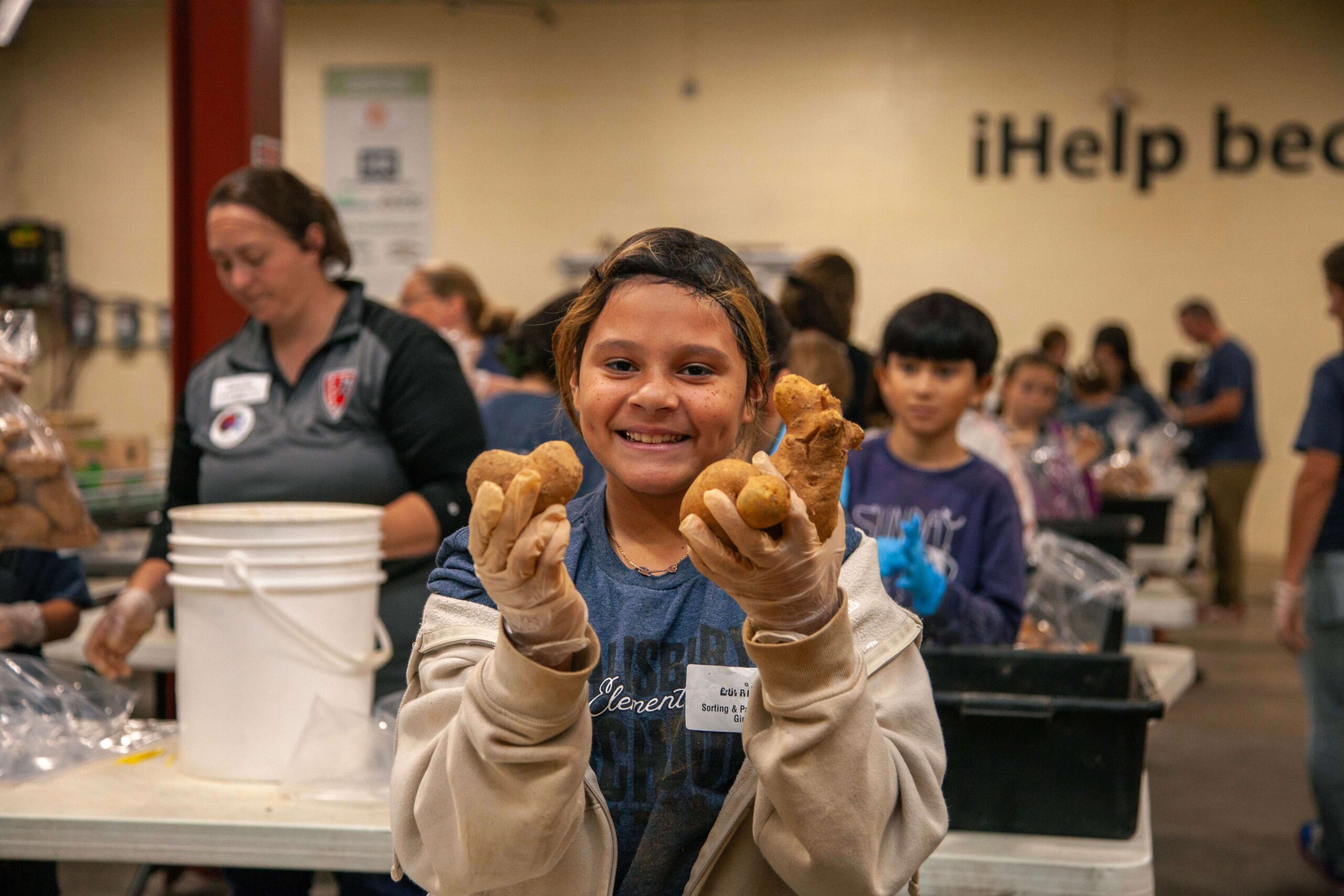 Smiling child holding potatoes while volunteering at a food distribution event, surrounded by other volunteers in a community service setting.
