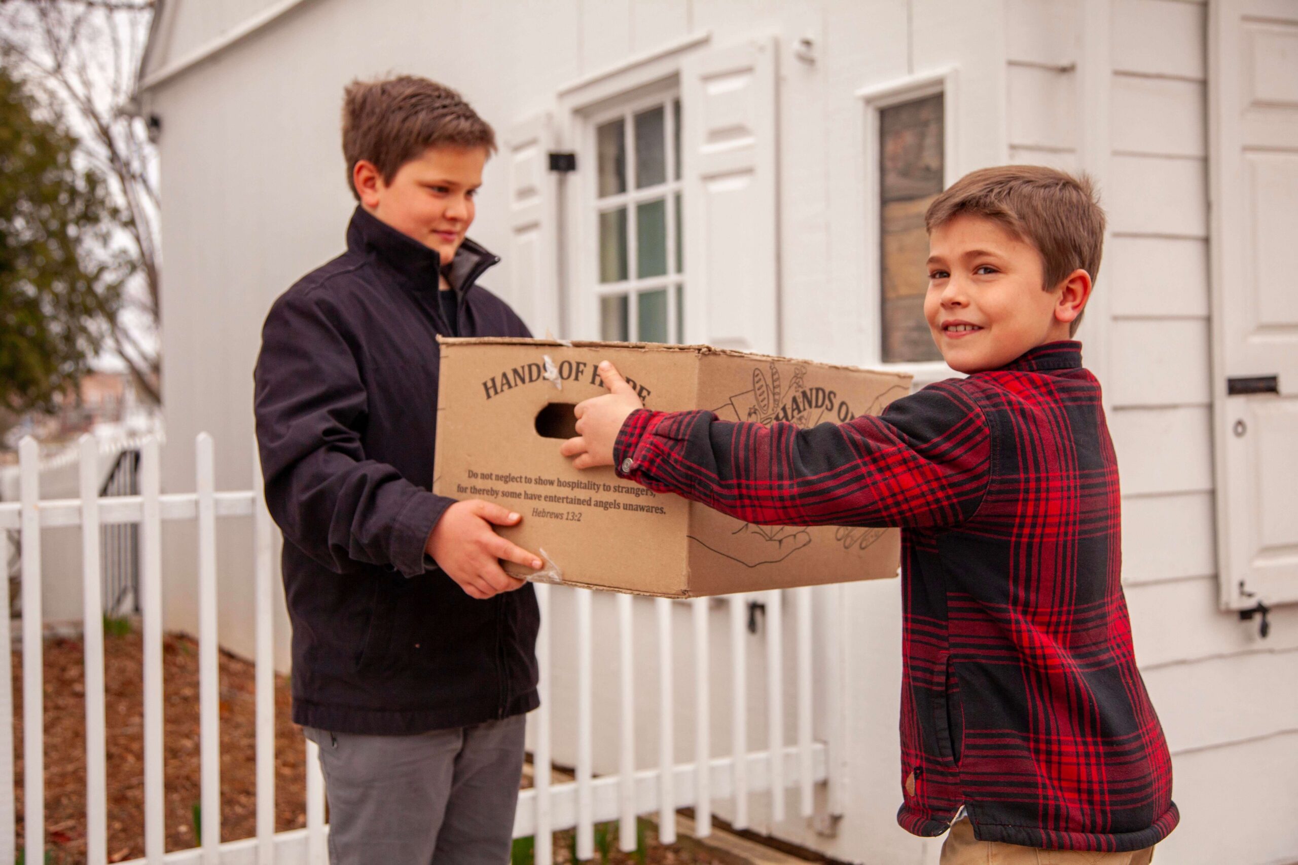 Two boys are participating in a charitable activity, handing over a box labeled "Hands of Hope." One boy, wearing a black jacket, is receiving the box, while the other, dressed in a red and black plaid shirt, is smiling as he passes it. The setting includes a white house and a picket fence, emphasizing community involvement and kindness.