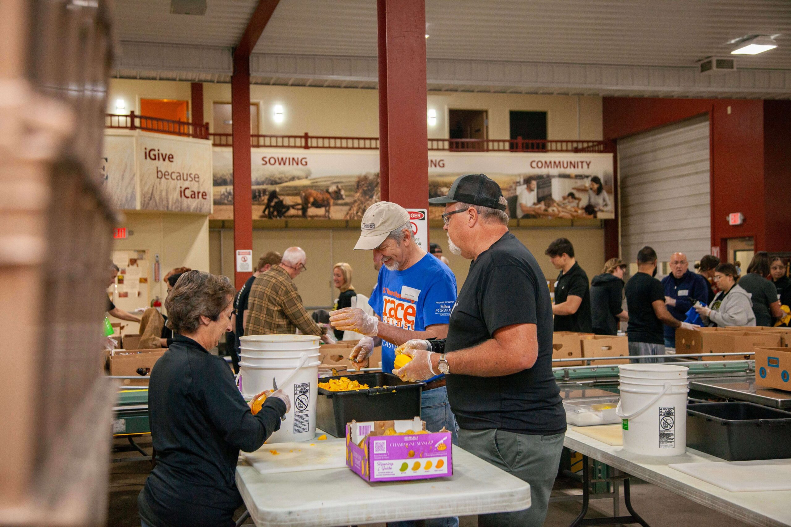 Volunteers participating in a community food packing event, sorting and preparing food items at tables in a large indoor space, with boxes and buckets visible in the background.