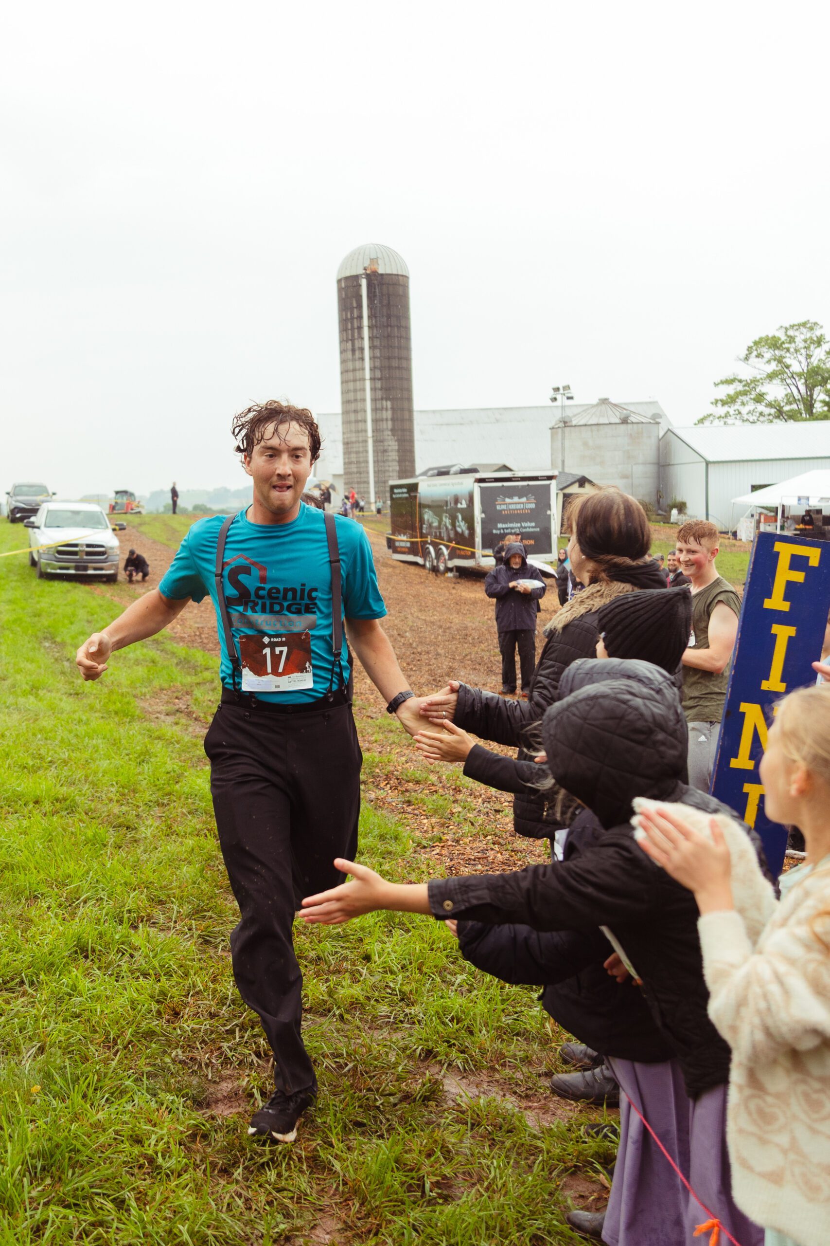 Runner in a blue Scenic Ridge shirt crosses the finish line at a rural event, greeted by children and spectators on a rainy day. A silos and farm buildings are visible in the background.