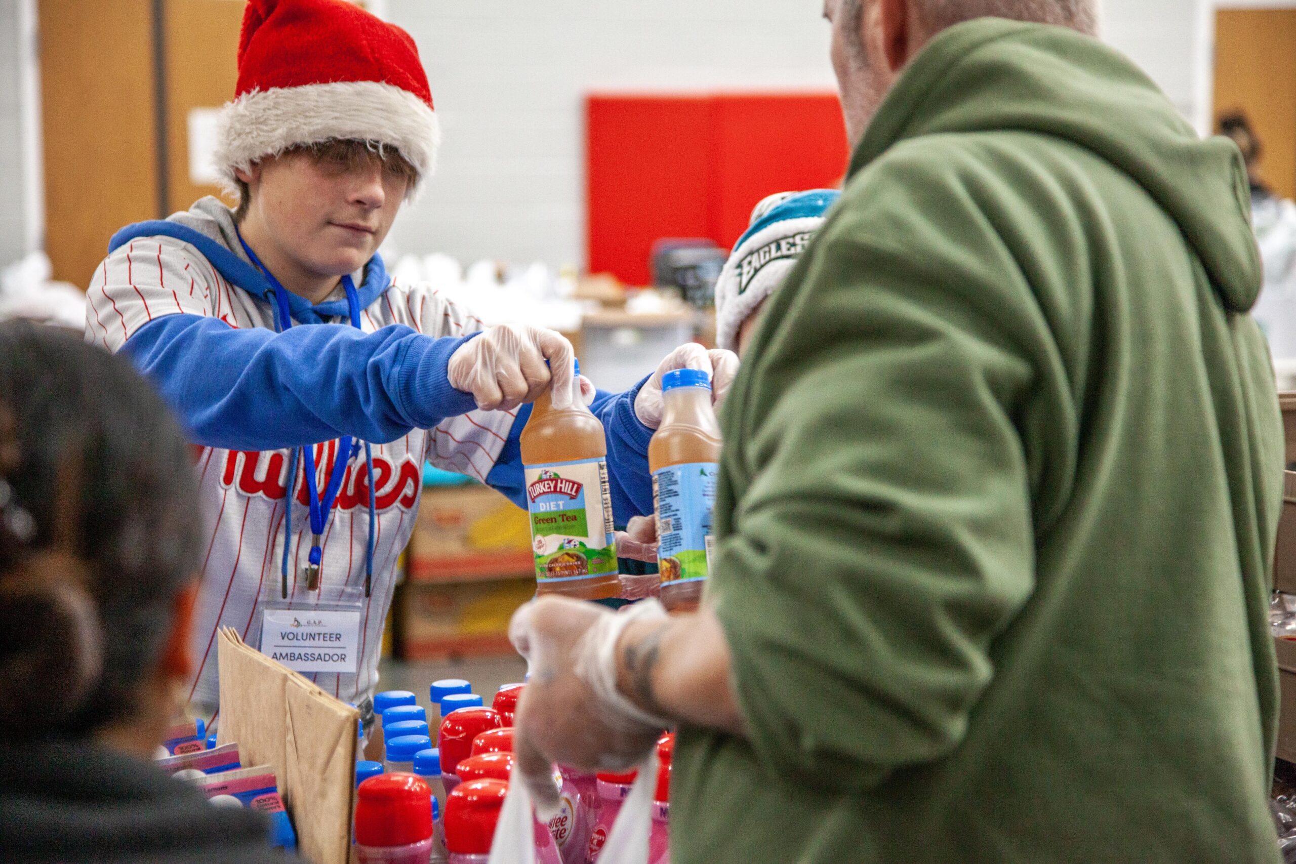 A young volunteer wearing a Santa hat hands bottles of iced tea to a client during a community food distribution event, showcasing the spirit of giving and support during the holiday season.