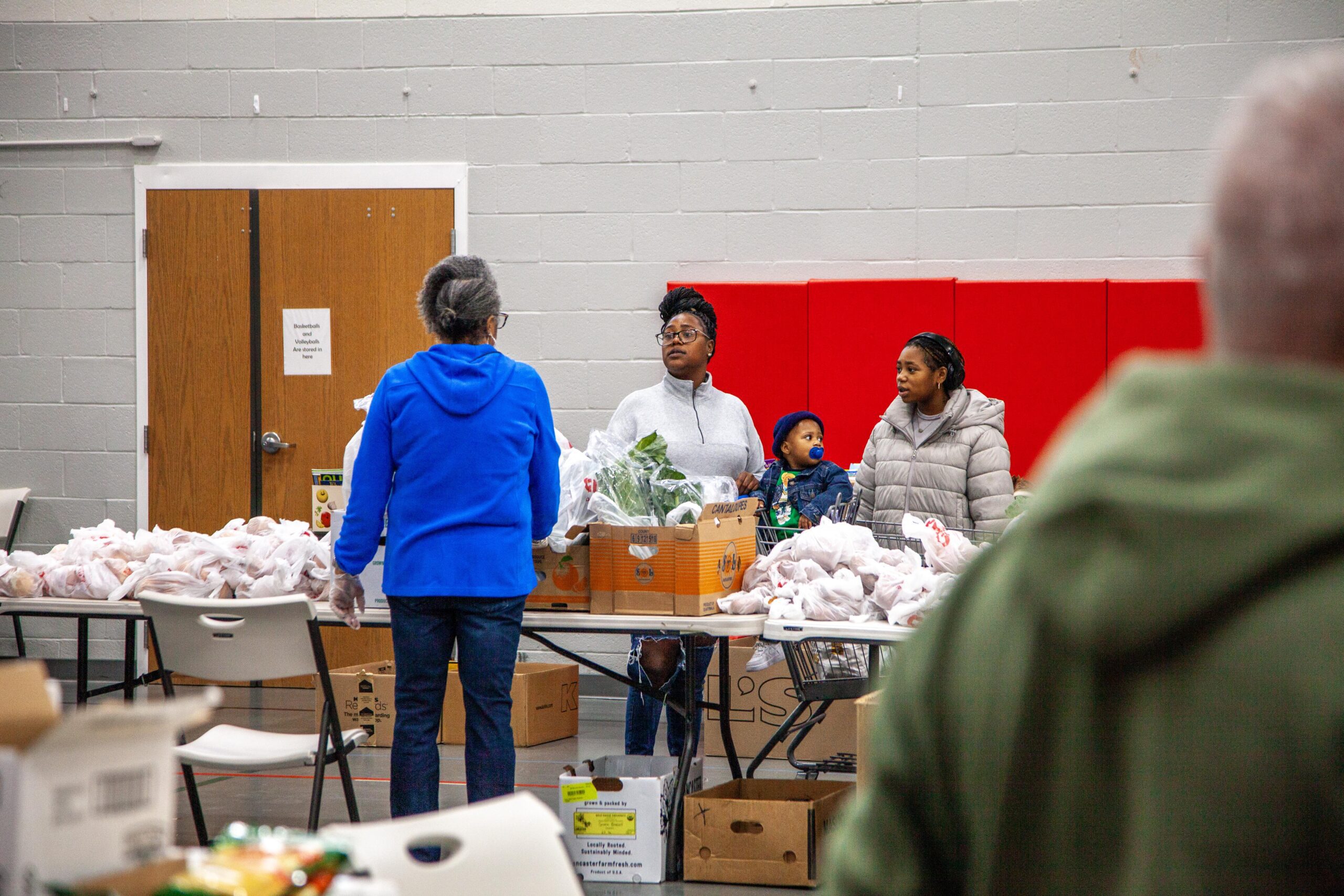 Community members gather at a food distribution event, interacting and selecting groceries from tables filled with bags and boxes of food in a communal setting.