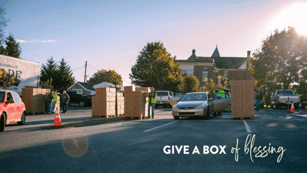 Community volunteers distribute food boxes from a truck labeled "Blessings of Hope" at a local event. Several vehicles are parked nearby, while people interact and organize the boxes in a sunny outdoor setting.