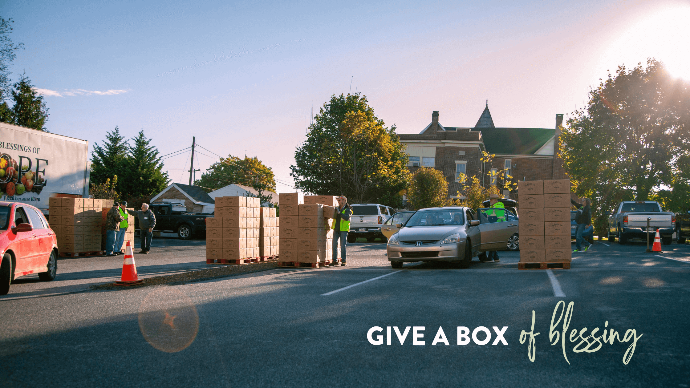 Community volunteers distribute food boxes from a truck labeled "Blessings of Hope" at a local event. Several vehicles are parked nearby, while people interact and organize the boxes in a sunny outdoor setting.