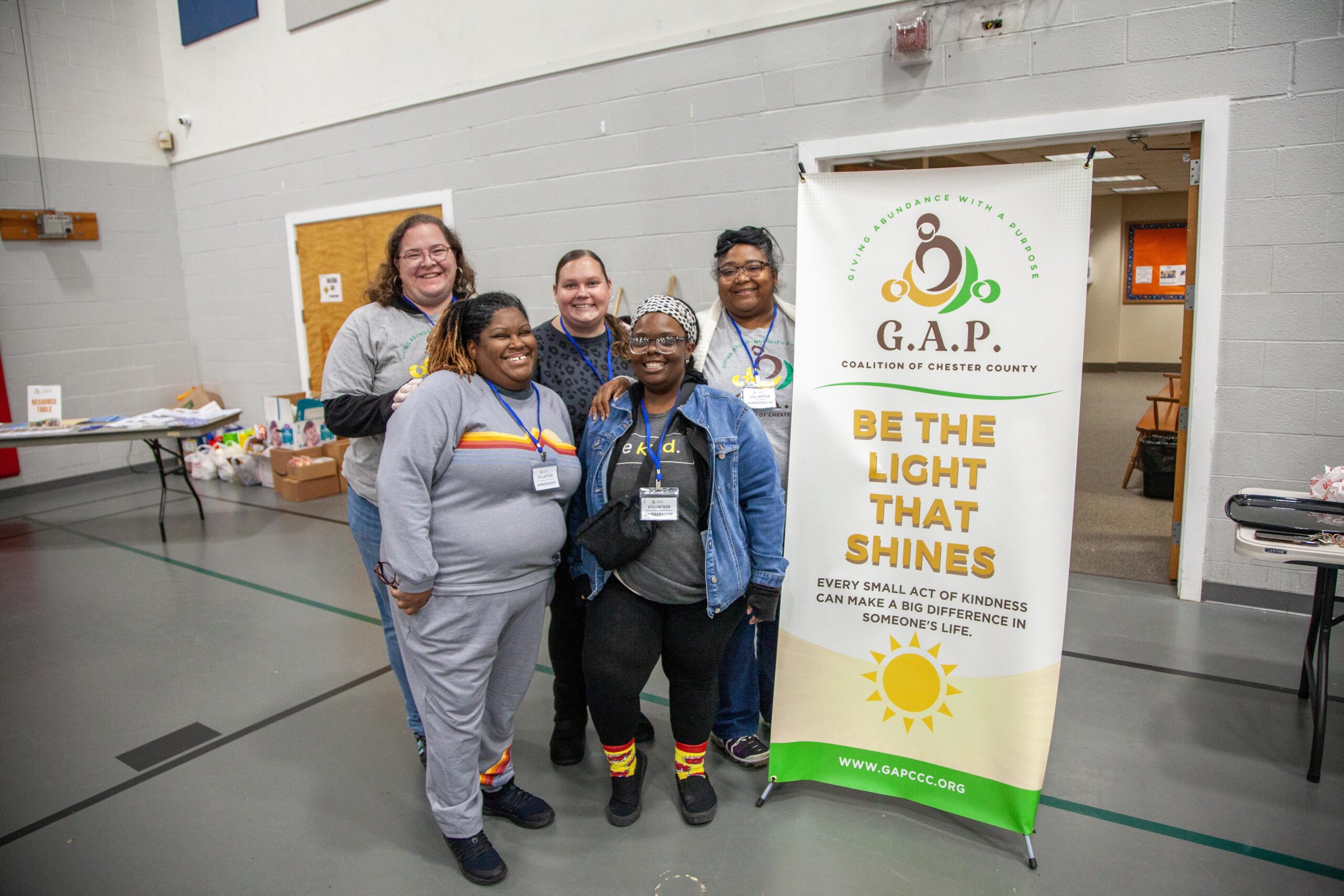 Group of five women smiling at a community event for the G.A.P. Coalition of Chester County, standing in front of a banner that promotes kindness and community support. The setting is indoors, with tables displaying supplies in the background.