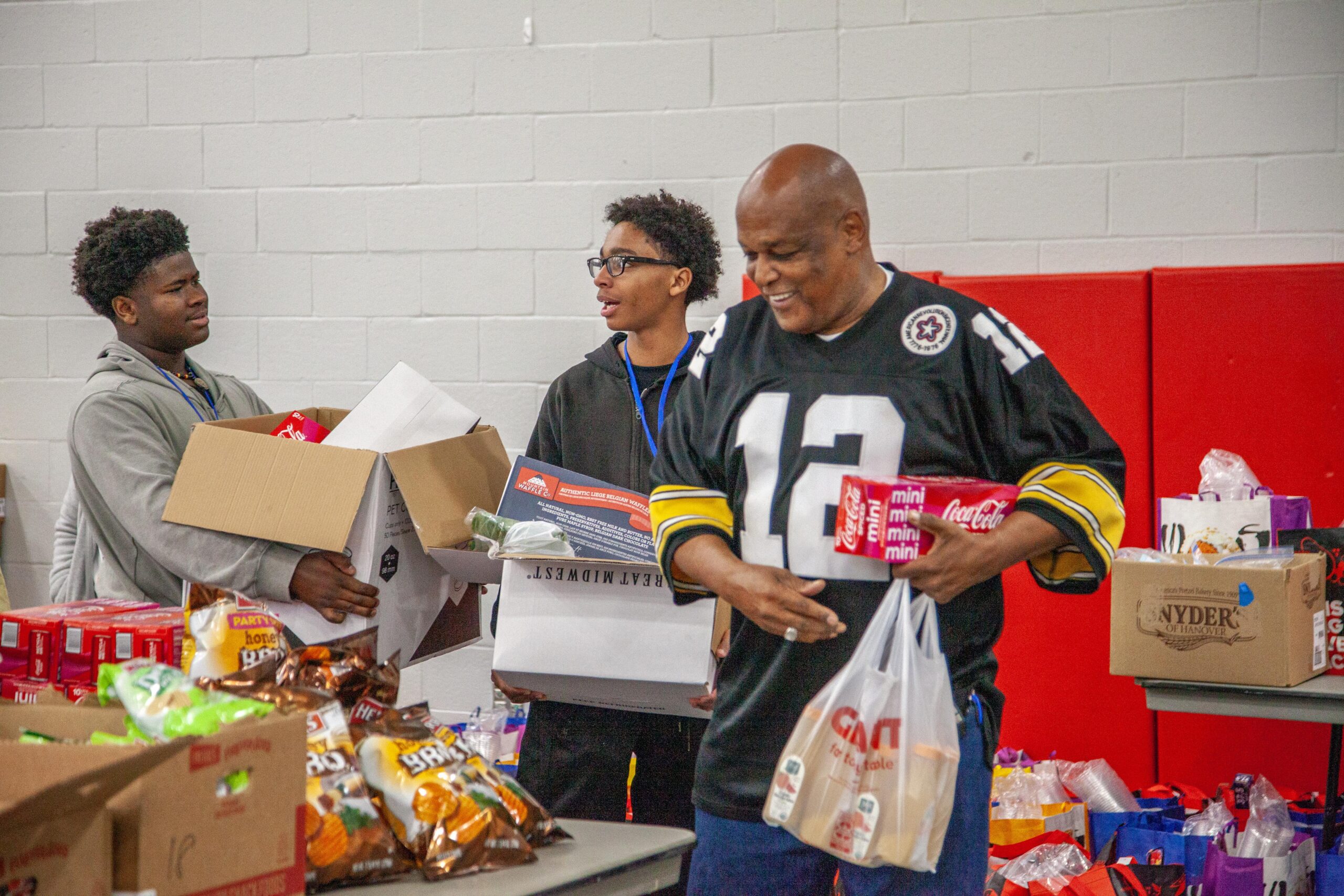 Group of three individuals engaged in a community food distribution event, featuring two young men carrying boxes and a smiling adult man in a sports jersey holding bags of snacks and beverages. Various food items are visible on tables, highlighting the effort to support the local community.