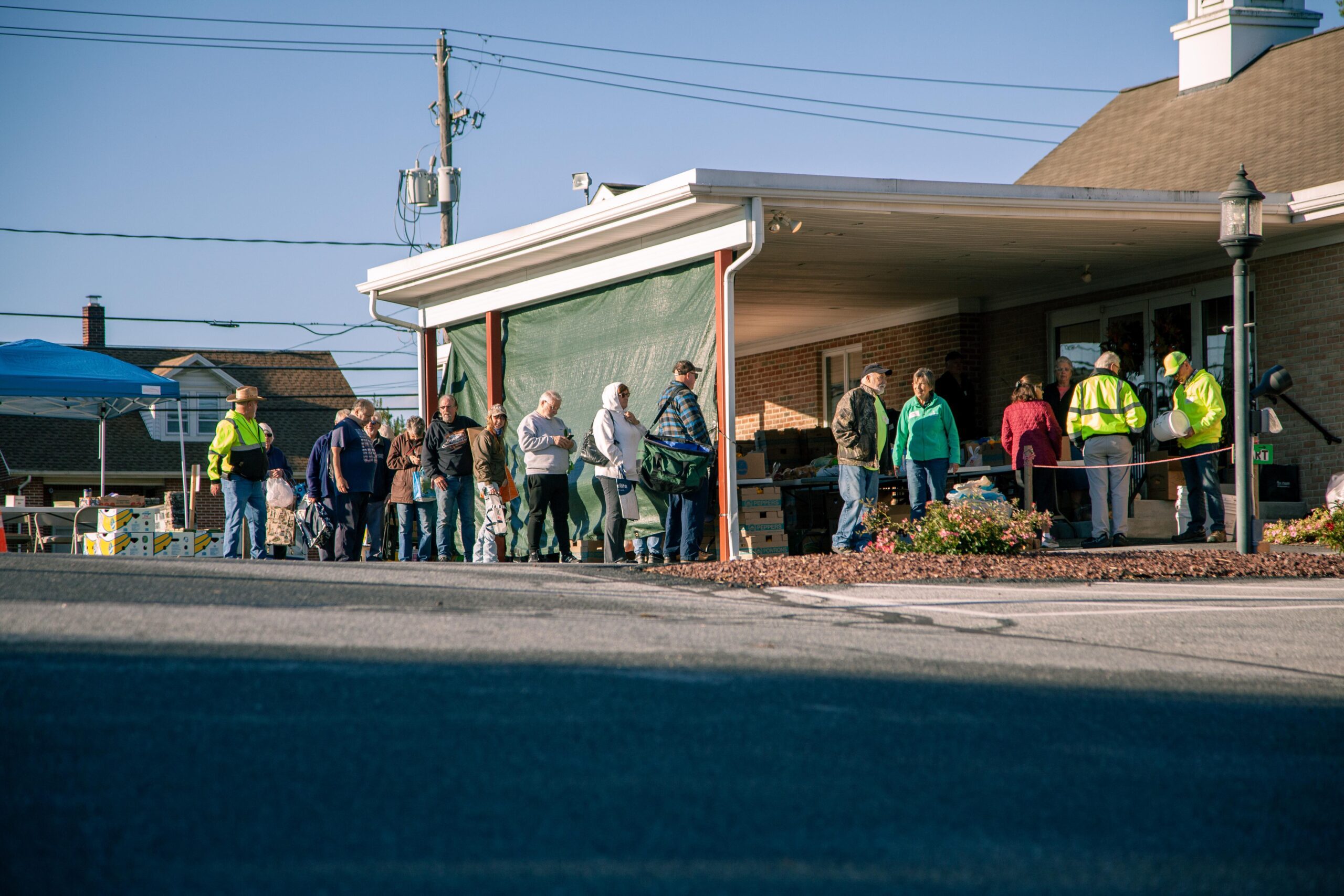 A diverse group of people standing in line outside a community center, waiting for assistance or services, with volunteers in bright vests interacting with them. The scene captures a sunny day, highlighting a supportive community environment.