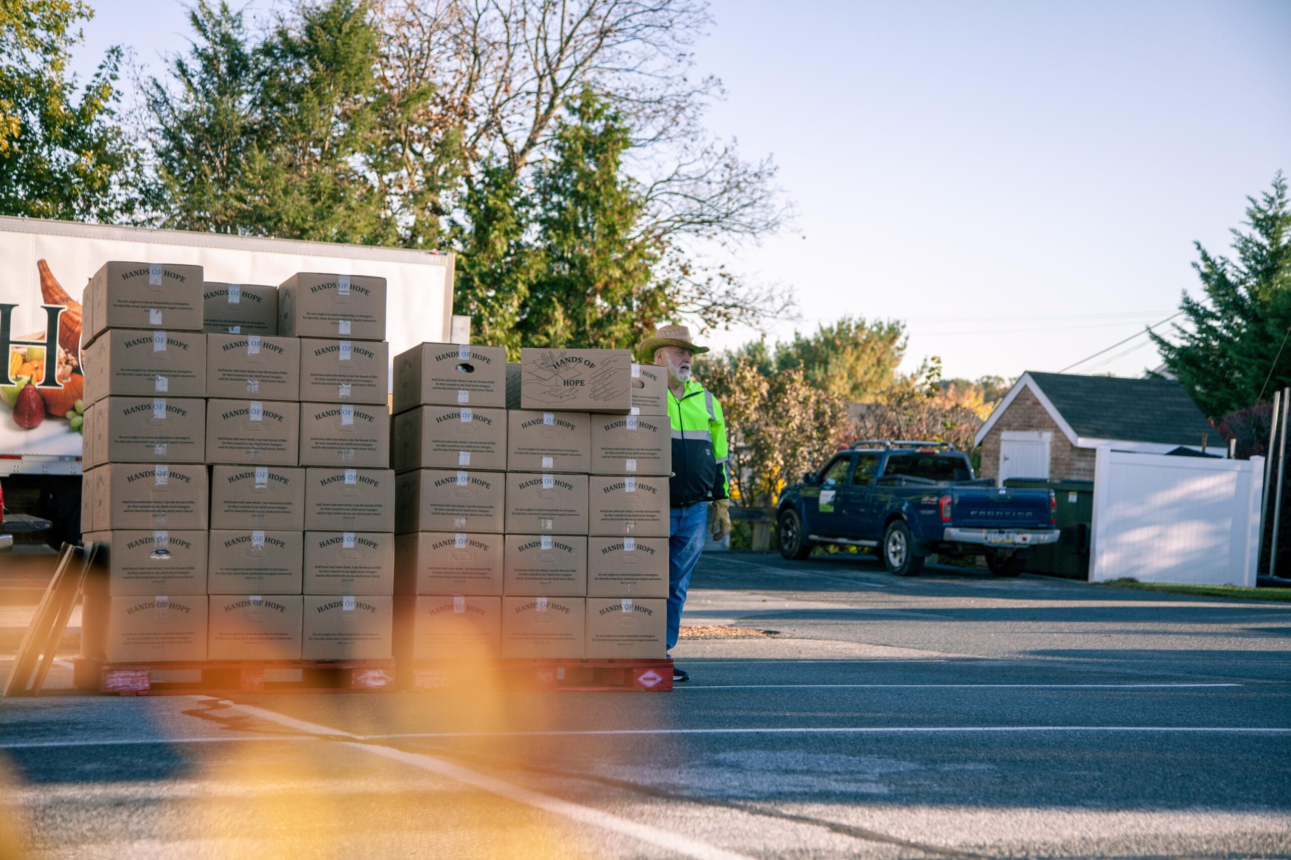 Man in safety gear overseeing stacked boxes labeled "Hands of Hope" at a distribution site, with a truck parked nearby and trees in the background.