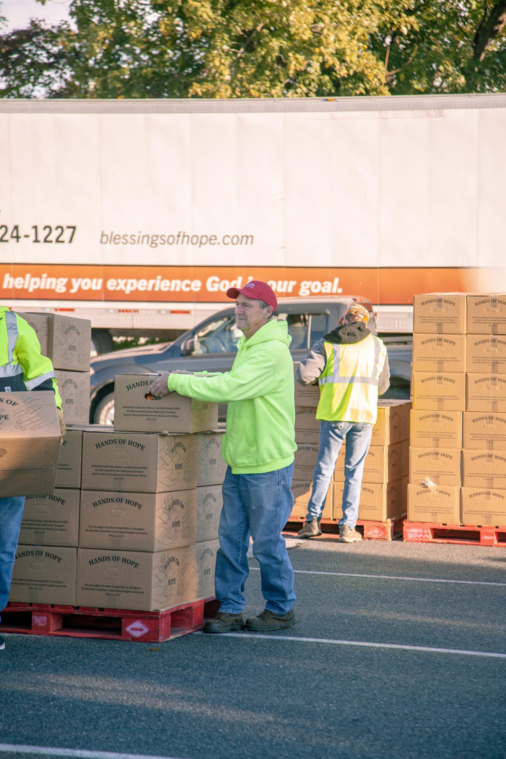 Volunteers loading boxes labeled "Hands of Hope" at a food distribution event, with a truck in the background promoting Blessings of Hope. The scene captures community engagement and support for those in need.
