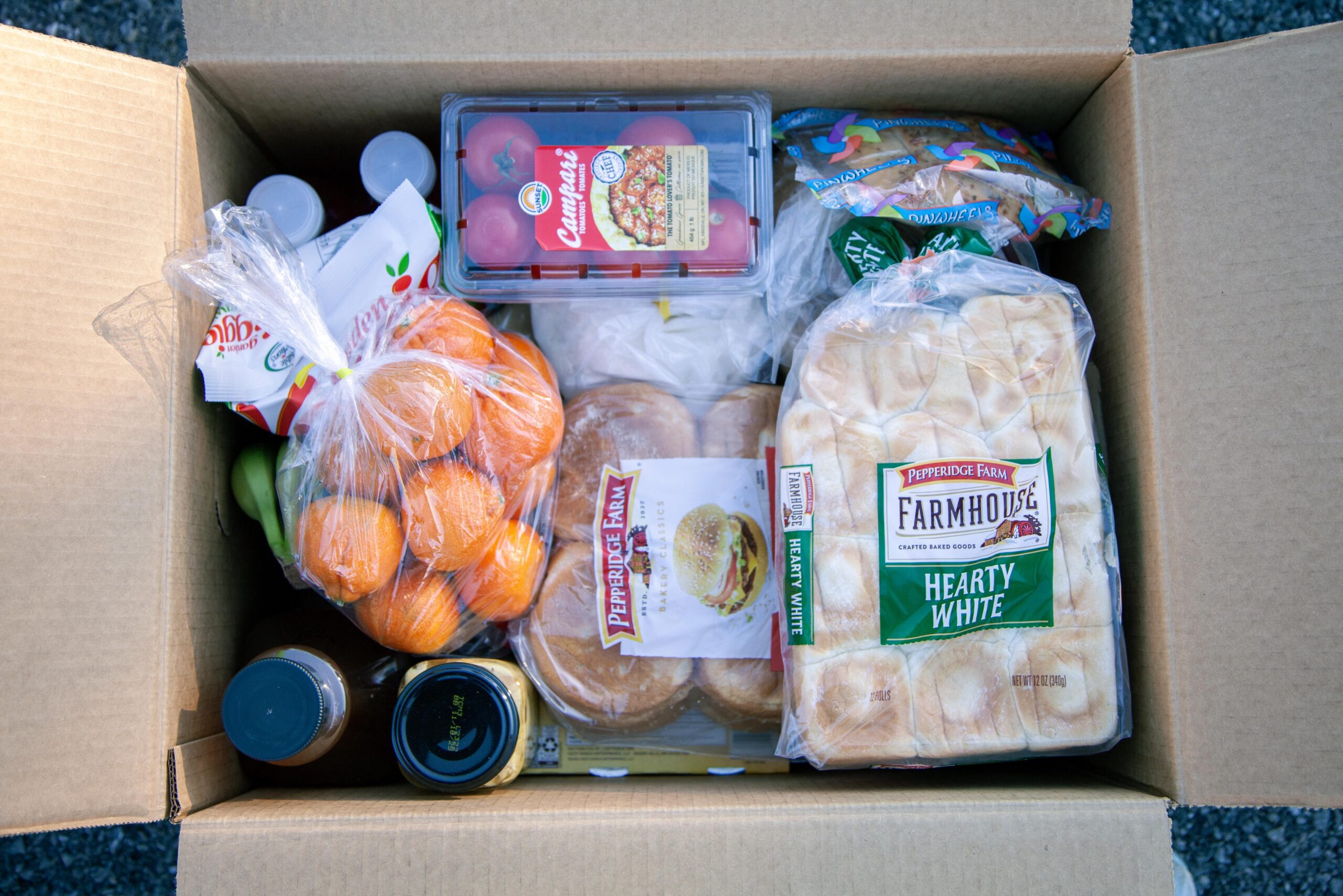 Cardboard box filled with assorted groceries including fresh fruits like oranges and bananas, packaged bread, tomato products, beverages, and condiments, highlighting a variety of essential food items for meal preparation.