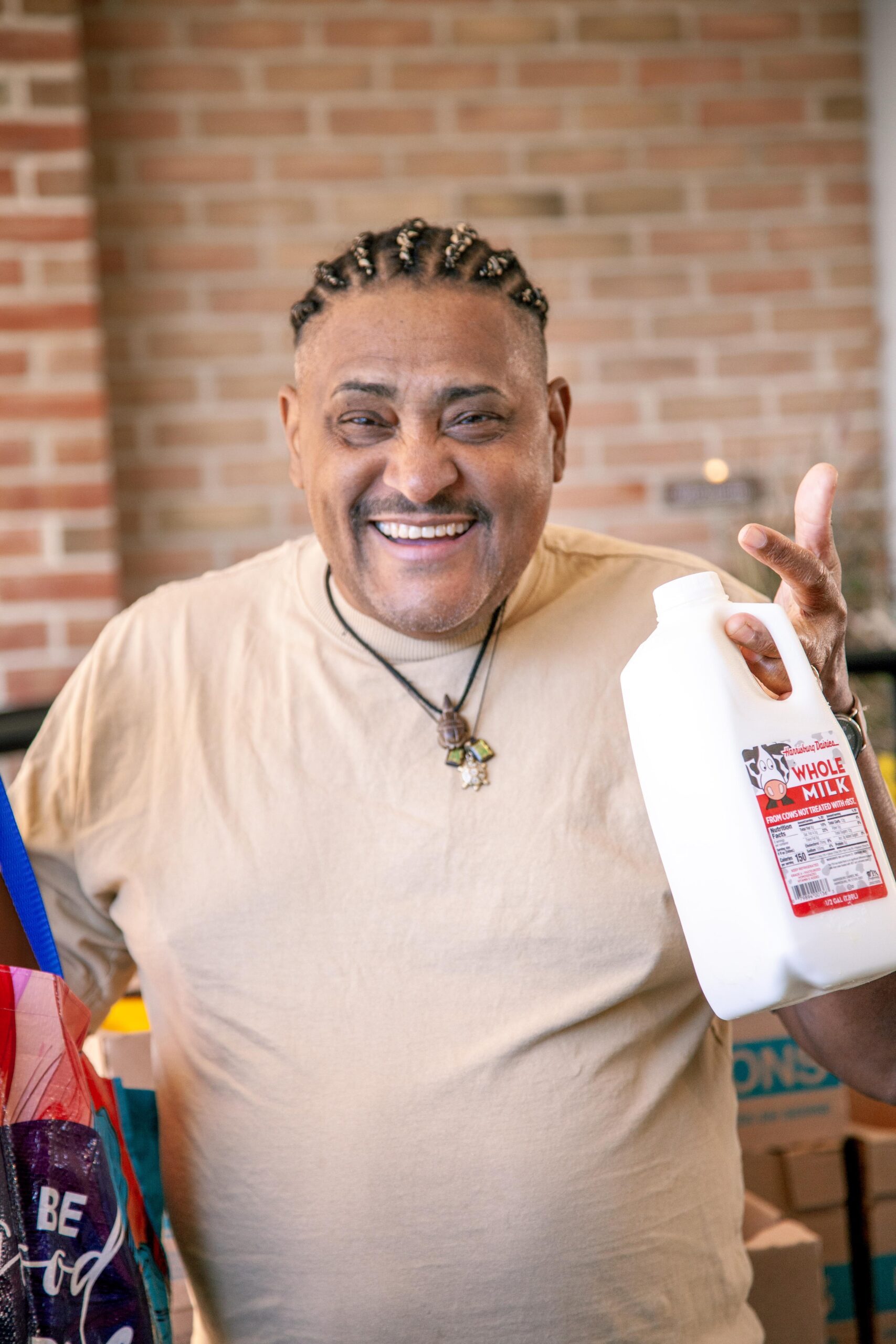 Smiling man holding a jug of whole milk while standing in a kitchen with a brick wall, wearing a beige shirt and a necklace, with grocery bags visible in the background.