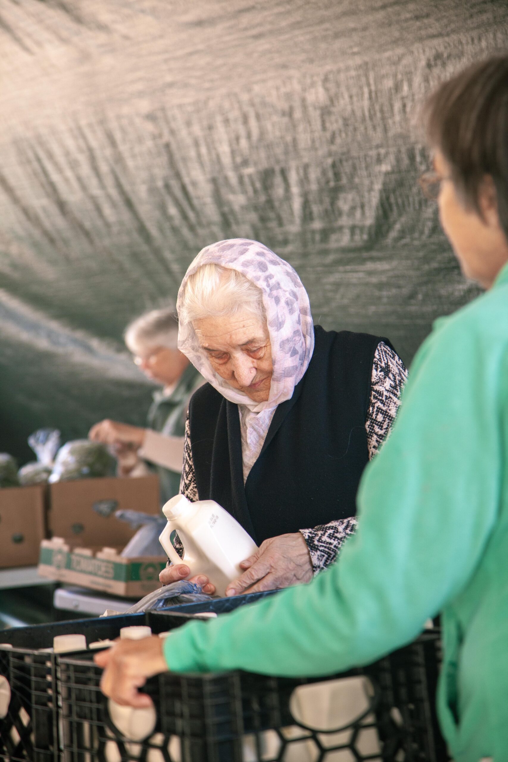 Elderly woman shopping at a market, holding a bottle while examining products. Another customer in a green sweater interacts nearby, with boxes of fresh produce visible in the background. The scene captures community engagement and the joy of shopping for local goods.
