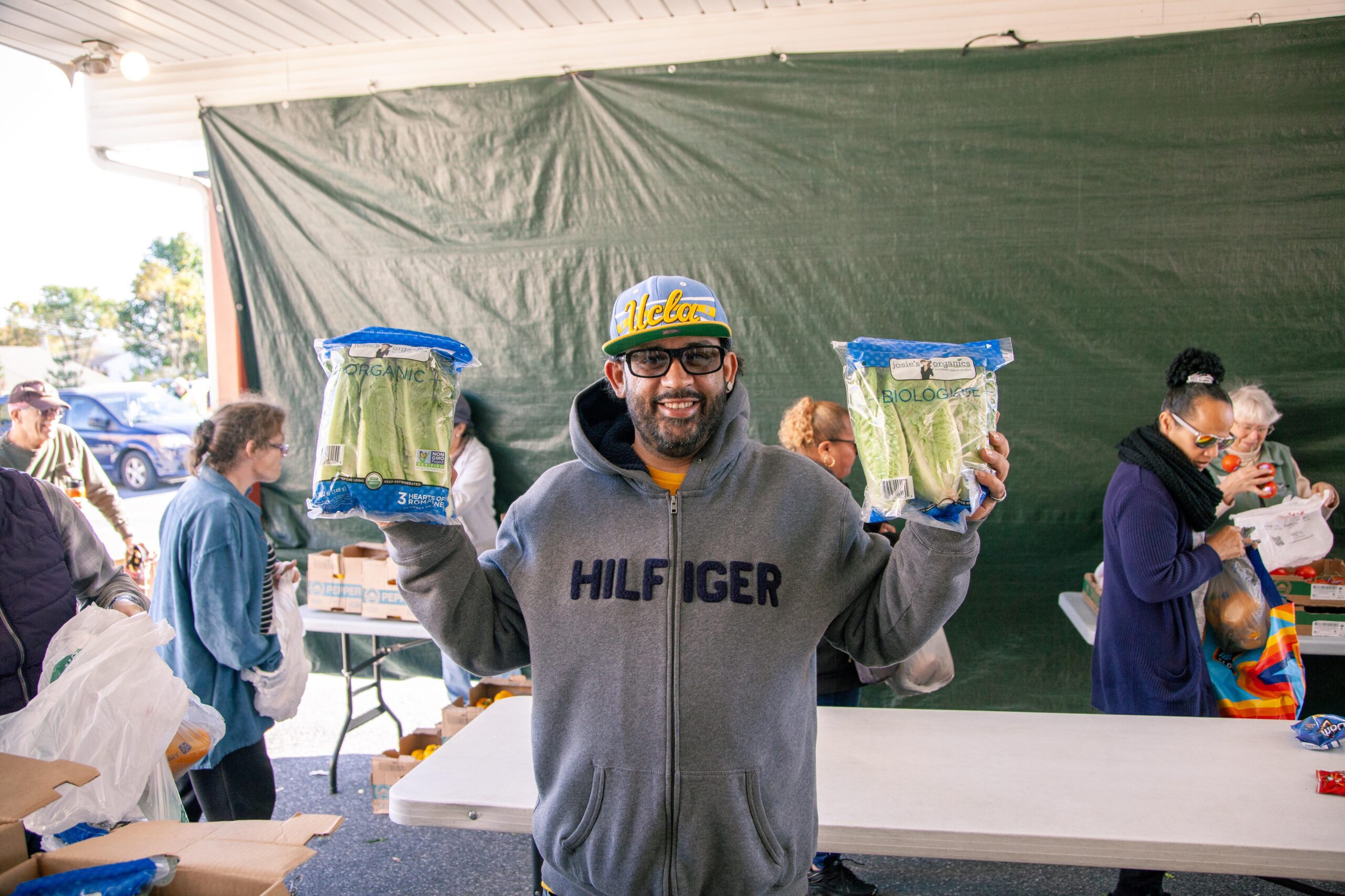 Smiling man holding two bags of organic celery at a food distribution event, surrounded by people collecting groceries.